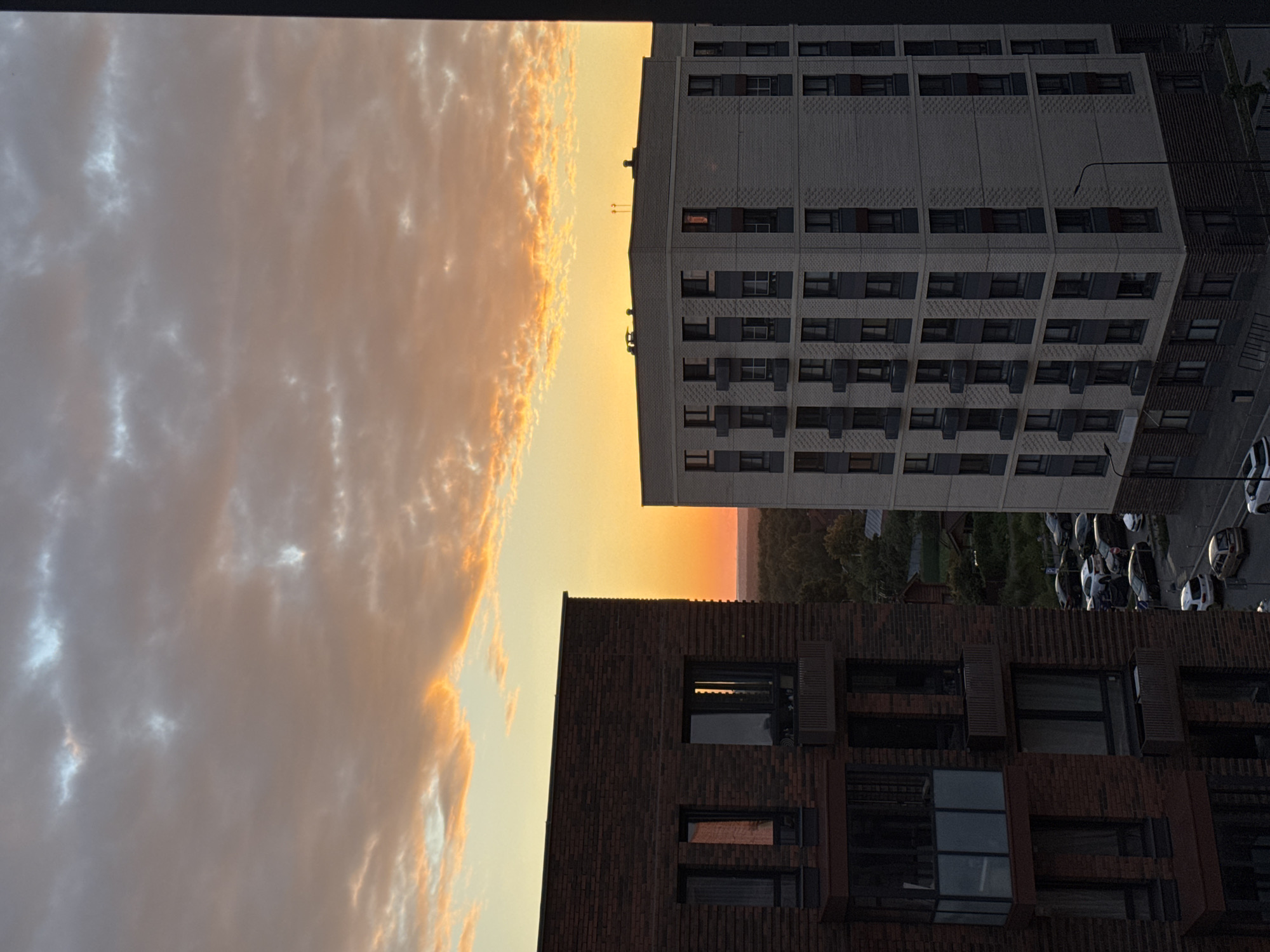 A view of an urban landscape during sunset, featuring two apartment buildings. The building on the left is constructed with dark red brick and has multiple windows and balconies. The building on the right is lighter in color, appearing to be made of light-colored bricks and concrete, also with numerous windows and balconies. The sky above is filled with clouds, tinged with orange and yellow from the setting sun. In the lower portion of the image, a street with several parked cars is visible. The scene is located in Laikovo, Russia.