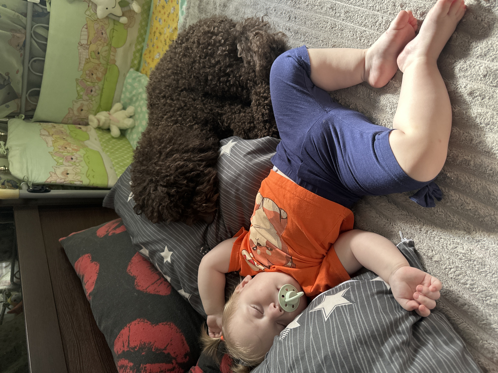 A young child sleeps peacefully on a bed with a brown dog lying nearby. The child, with blonde hair in pigtails and a pacifier in their mouth, is wearing an orange t-shirt with a cartoon animal print and blue shorts. They are lying on their back on several patterned pillows, with their legs extended. The dog, a curly-haired breed, is curled up and appears to be sleeping as well. The scene is likely indoors, given the presence of pillows and blankets, and the time of day is suggested to be daytime by the soft lighting. The overall mood is one of calm and comfort. The pillows have various designs, including one with a red lips pattern and another with gray stripes and white stars. The background includes more pillows with cartoon animal motifs and a yellow patterned fabric. No visible text is present in the image. The location context indicates this scene is taking place in Sayanogorsk, Russia.