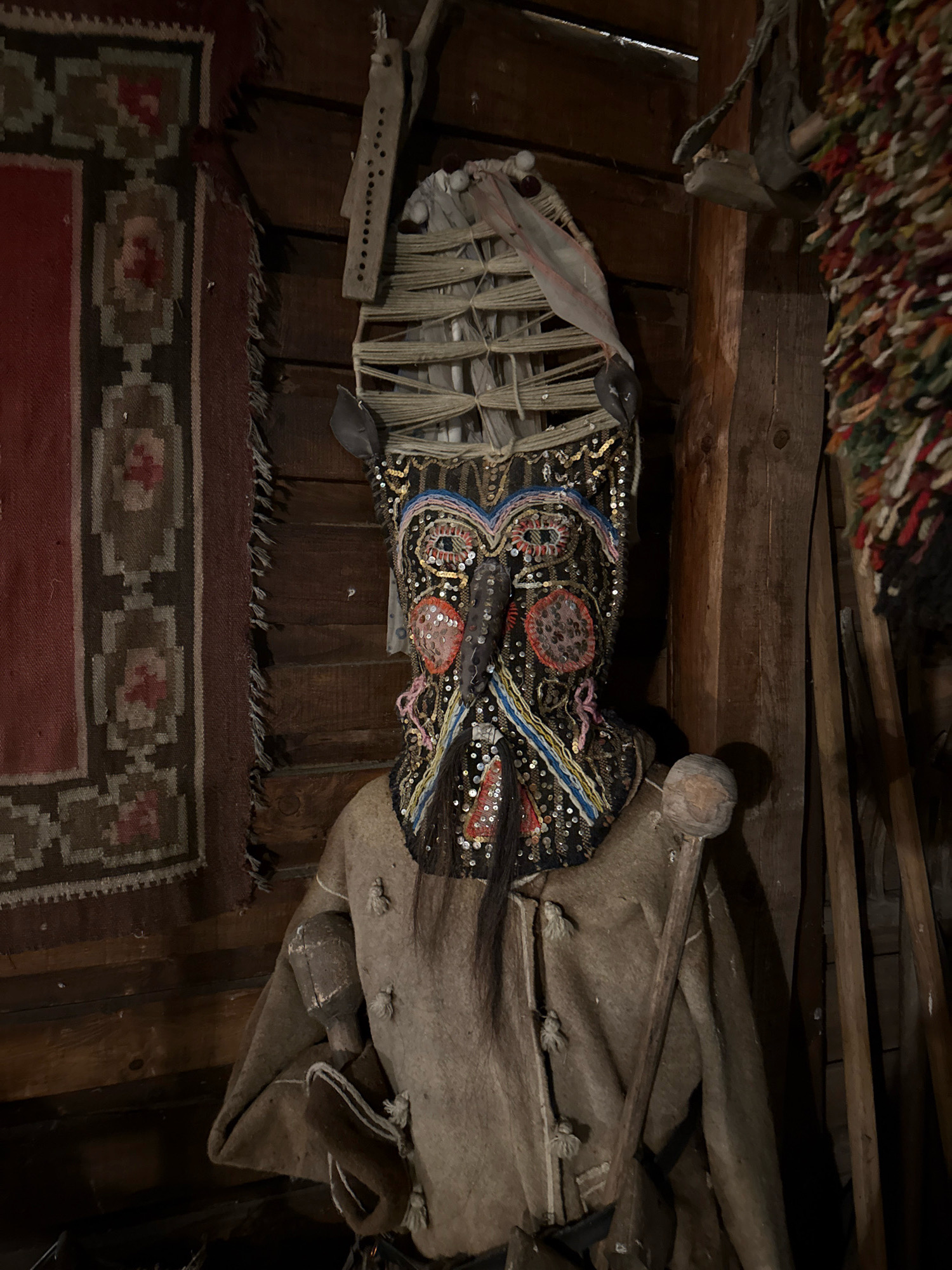 The image shows a wooden structure, likely a room in a traditional Bulgarian house in Chernevo, Bulgaria. A figure, possibly a mannequin, is dressed in a rustic, earth-toned garment and wears an elaborate, decorated mask. The mask is adorned with sequins, beads, and various colorful elements, creating a striking and somewhat unsettling appearance. Behind the figure, a woven rug with geometric patterns hangs on the wooden wall, contributing to the traditional aesthetic of the space. Several wooden objects, including what appears to be a staff or tool, are visible in the foreground and to the right of the figure, leaning against the wooden walls. The overall impression is one of cultural heritage preservation, showcasing traditional attire and possibly ritualistic objects within a rustic interior setting.
