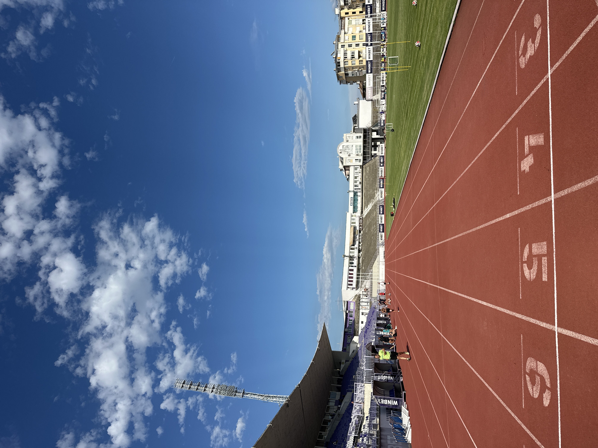 A low-angle, wide shot captures a red running track in a stadium with the stands on the left and the grass field on the right. The track has white lane markings and numbers indicating lanes 3, 4, 5, and 6 are visible in the foreground. Several people are visible on the track, some running and some standing near the bleachers. The stadium is situated in Veliko Tarnovo, Bulgaria, indicated by the "WINBET" advertisements around the stadium. In the background, buildings with yellow and white facades are visible. The sky is blue with scattered white clouds. A tall light tower is positioned to the left of the stadium.