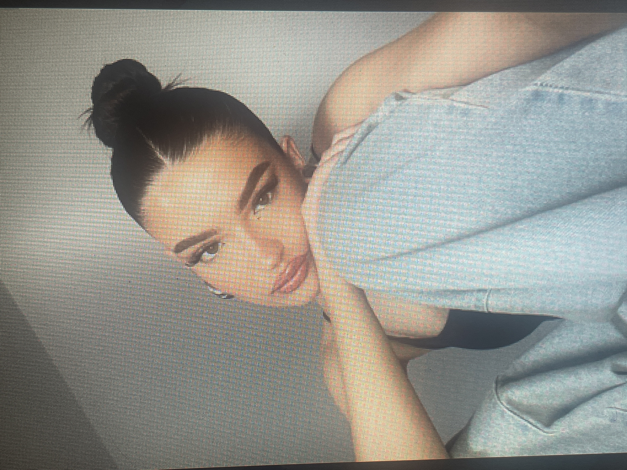 The image shows a close-up portrait of a young woman with her hair pulled back in a bun. She is leaning her head on her knee, and her gaze is directed towards the viewer. She is wearing a dark top and light blue jeans. The background is a neutral, textured wall. The lighting suggests an indoor setting, likely a studio or a well-lit room. The image quality is pixelated, suggesting it might be a photograph taken from a screen or a low-resolution digital capture. There is no indication of the city of Mantov, Czechia, in this image.