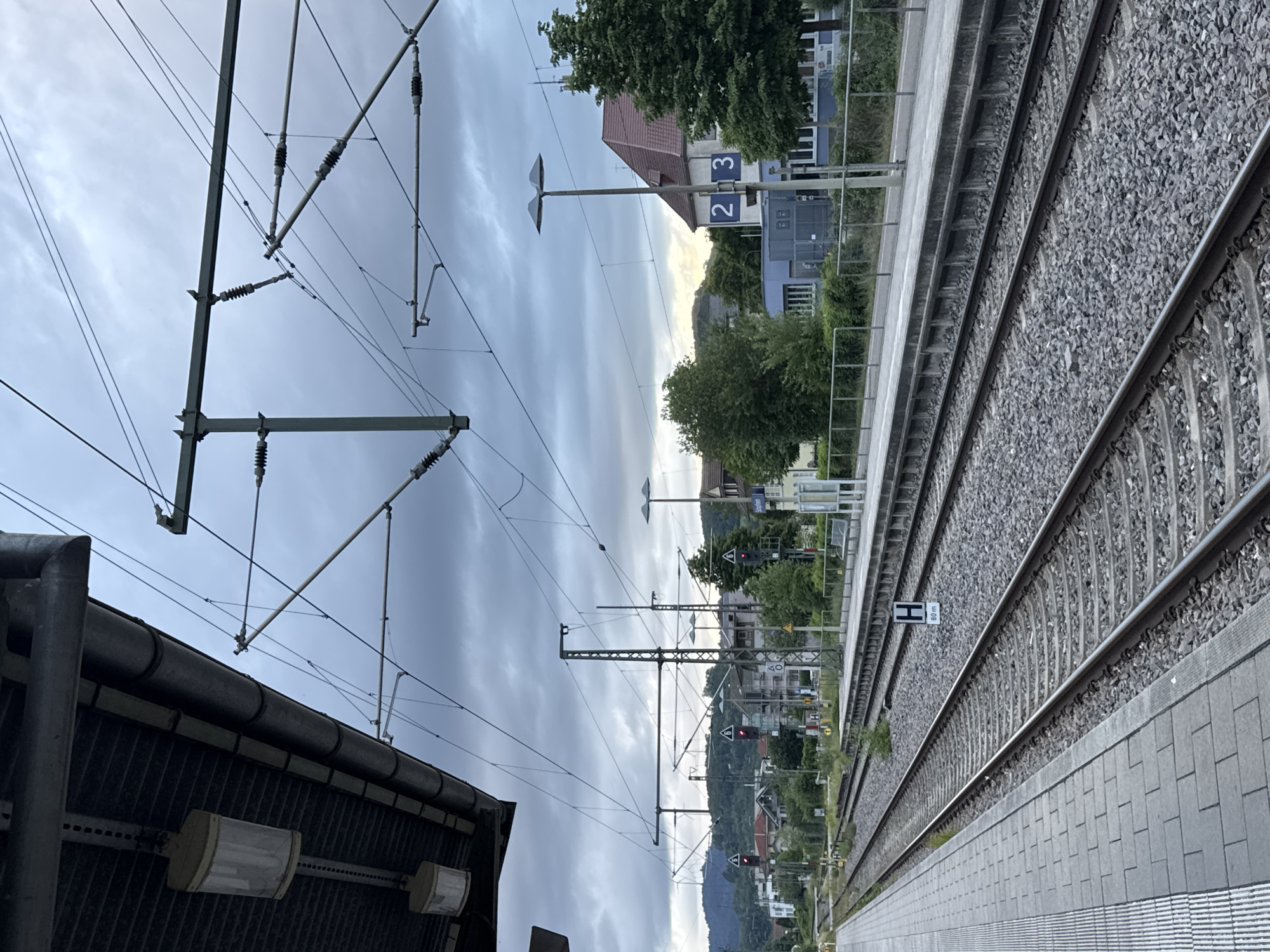 An eye-level, medium shot captures a railway station on an overcast day in Schopfheim, Germany. The foreground is dominated by a gray, tiled train platform on the left and parallel train tracks on the right. The tracks are surrounded by gravel and extend into the distance. Overhead, a complex network of electrical wires and support structures for the train system crisscrosses the frame.
In the middle ground, more train tracks are visible, along with a concrete platform. To the right of the tracks, a sign displaying "2 3" is affixed to a lamppost. Beyond the platform, a low-rise building with a red-tiled roof and light-colored walls stands, partially obscured by trees. Further in the background, more buildings and trees are scattered across a hilly landscape. A signal light showing red is visible on a pole to the left. The sky is a muted gray, indicating cloudy conditions.