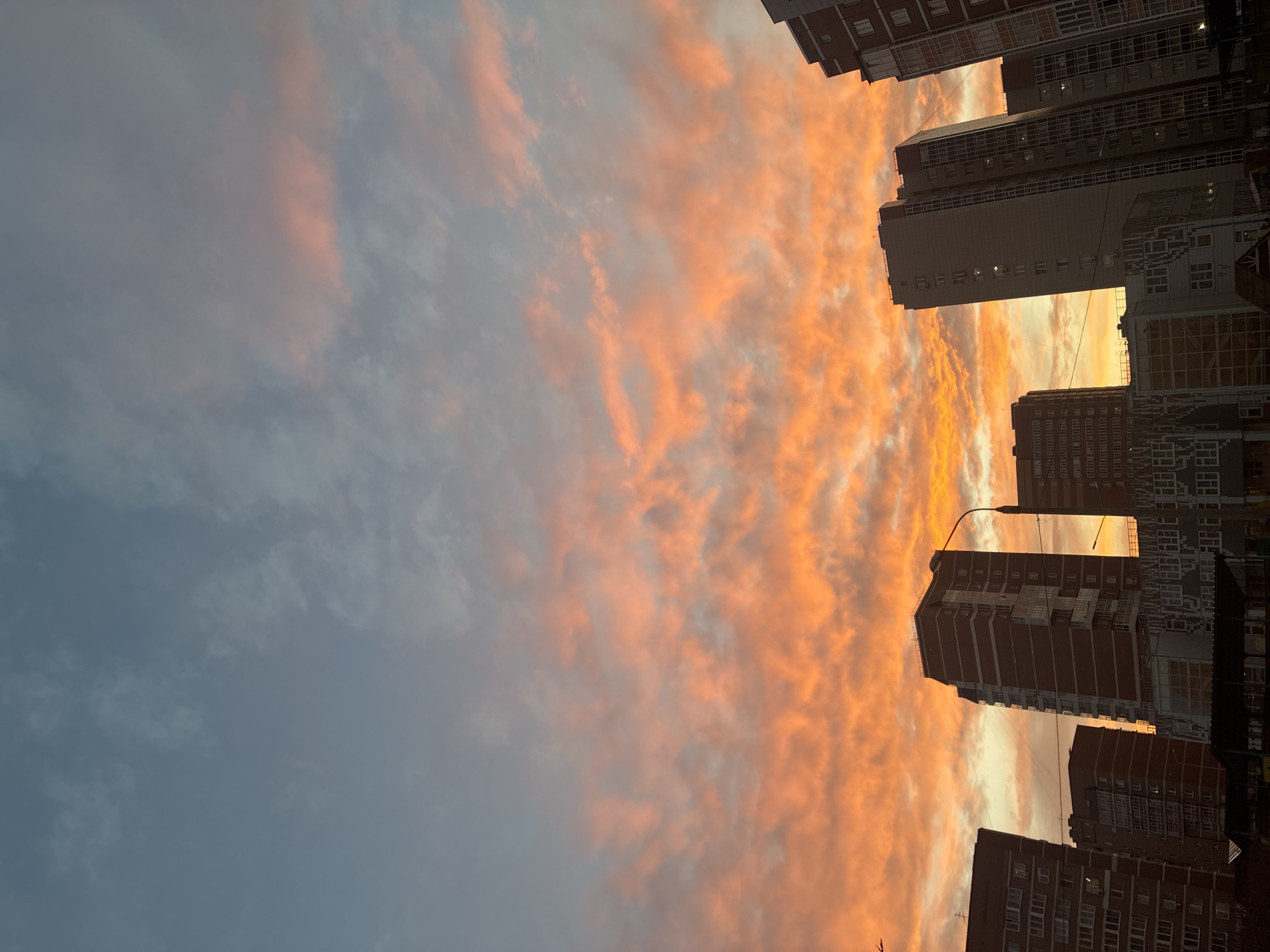 The image displays an urban or suburban landscape dominated by a vividly colored sky at what appears to be either sunrise or sunset. The sky, occupying the majority of the frame, is filled with dramatic, streaky clouds painted in shades of intense orange, fiery red, and soft pink, transitioning to a lighter blue-grey towards the upper left. These warm-toned clouds create a striking contrast with the darker, cooler tones of the silhouetted buildings below and to the right.
Several tall, multi-story buildings are visible, appearing as dark masses against the luminous sky. Their architectural details are somewhat obscured by the backlight, but they seem to be residential or commercial structures with numerous windows. A prominent street light pole with a lamp is visible near the center-right, positioned between two of the taller buildings. Faint lines, possibly utility wires, can be seen crisscrossing the sky in some areas. The scene captures a moment of natural beauty as the sky transitions colors, highlighting the verticality of the urban structures against the vast expanse of the sky. No human activity or visible text is discernable in the image.