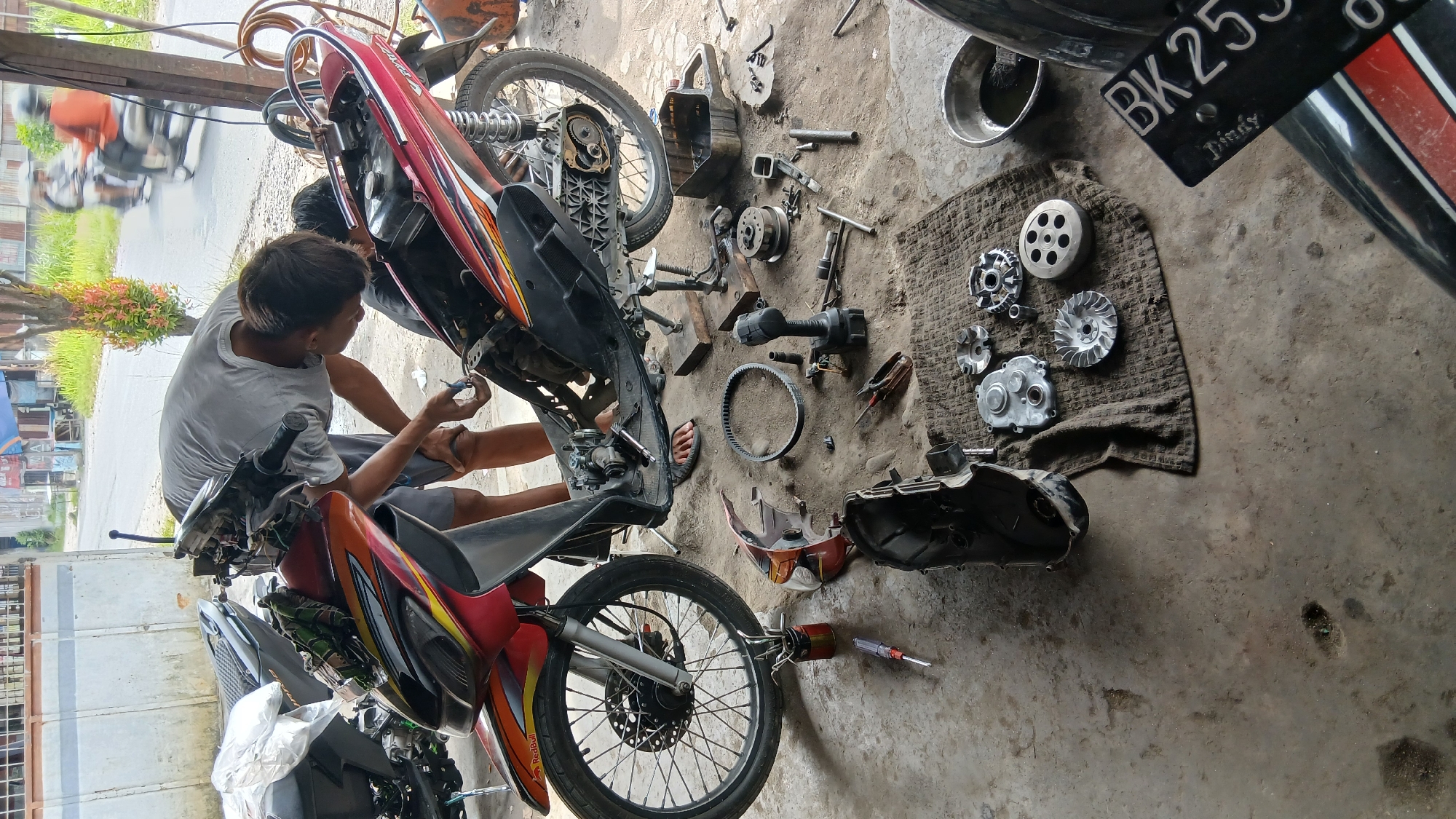 In Klambir Lima Kebun, Indonesia, a mechanic is working on a red and black scooter in what appears to be an outdoor workshop, possibly a residential area. The scooter is partially disassembled, with its engine cover removed and various parts, including a belt and clutch components, laid out on the ground. The mechanic, wearing a grey t-shirt and shorts, is focused on the scooter's engine. Another person is partially visible behind the scooter, also seemingly involved in the repair. The ground is concrete, with some dirt and oil stains. Tools, including wrenches, pliers, and a drill, are scattered around the work area. In the background, a street with a few passing motorcycles and a building can be seen, suggesting a semi-urban environment. A license plate with "BK 255" is visible in the foreground.