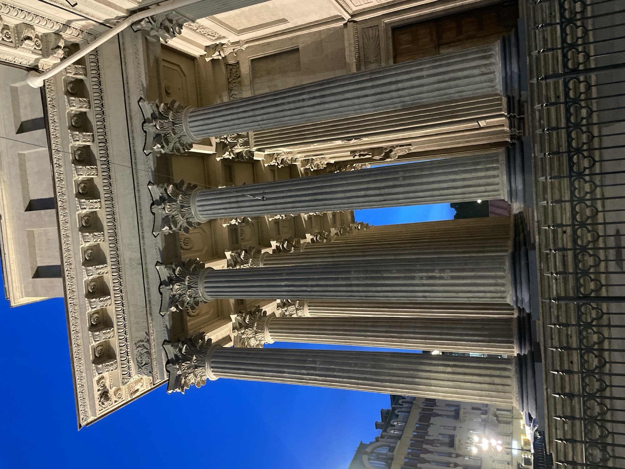 The image displays a low-angle, upward-facing view of the Kazan Cathedral in Saint Petersburg, Russia, during twilight or early night. In the immediate foreground, a black, ornate iron fence with repeating trefoil heart-shaped motifs runs horizontally. Beyond the fence, a series of imposing, fluted columns with elaborate Corinthian capitals rise vertically, illuminated by artificial light. These columns, appearing grey-beige, are supported by dark, circular bases resting on a series of grey stone steps. Above the columns, a richly detailed entablature features a cornice with dentils, egg-and-dart molding, and floral friezes including rosettes. The upper part of the building's facade, including a plain wall section and a wooden door on the far right, is visible. The sky, visible through and above the columns, is a deep, clear blue. To the far left, behind the cathedral's colonnade, a multi-story building with arched windows and a complex roofline is partially visible, along with distant streetlights. No individuals are present in the scene.