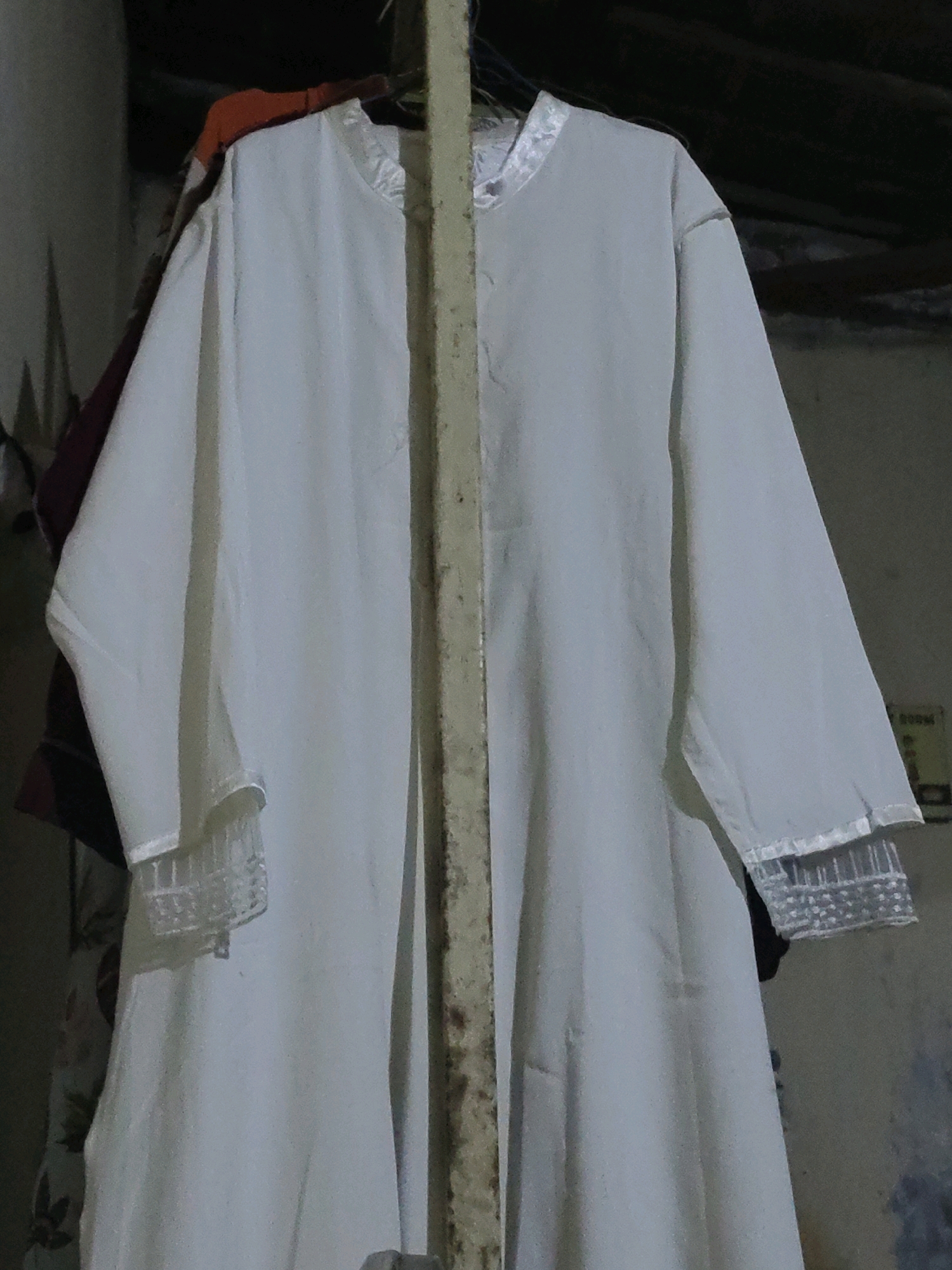 A white garment, possibly a dress or a robe, is hung on a vertical pole in an indoor setting. The garment is made of a lightweight fabric and features long sleeves with decorative cuffs. The collar of the garment also appears to have some embellishment. The background is dimly lit and appears to be a somewhat weathered wall. There are other dark-colored garments visible behind the white one, also hanging from a support. The scene suggests a domestic or storage area. The image was captured in Bulak Lor, Indonesia.