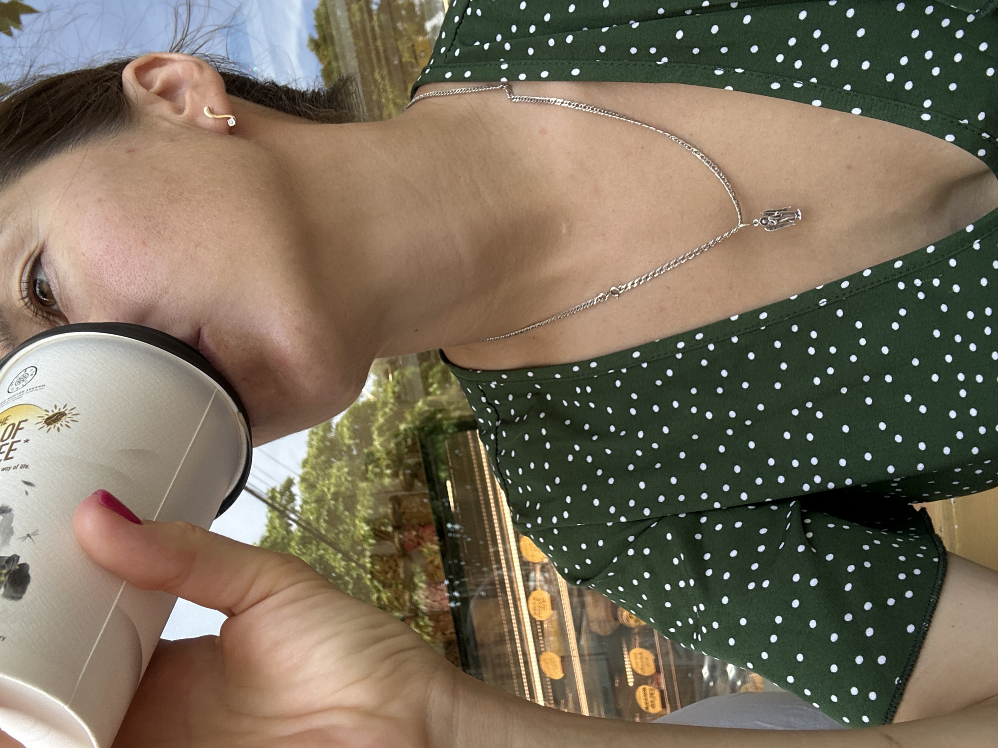 A close-up, waist-up shot shows a person holding a paper cup to their lips and drinking. The person is wearing a green dress with white polka dots and a silver chain necklace with a small, detailed pendant. They have a small, musical note-shaped earring with a tiny diamond on their right ear. The background appears to be a cafe or bakery, with display cases filled with pastries visible through a window. The environment outside the window suggests it is daytime, with trees and a blue sky. The image was taken in Nha Trang Ward, Vietnam.