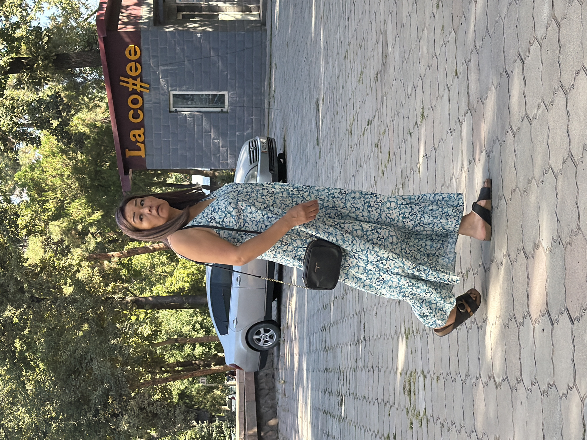 A medium shot captures a woman walking on a stone-tiled path in an outdoor setting, possibly a park or pedestrian area. She is wearing a blue and white floral print maxi dress and dark sandals. A small black crossbody bag with a gold chain strap is slung over her right shoulder. She has dark hair, pulled back, and is looking towards the left of the frame with a neutral expression.
In the background, a silver sedan is parked on the right side of the image. To the right of the car, a small building with a dark red roof has the word "La co##ee" displayed in yellow lettering. The building appears to be a kiosk or a small cafe. Trees with green foliage provide shade in the background, indicating a sunny day. The ground is paved with light gray, interlocking stone tiles.
