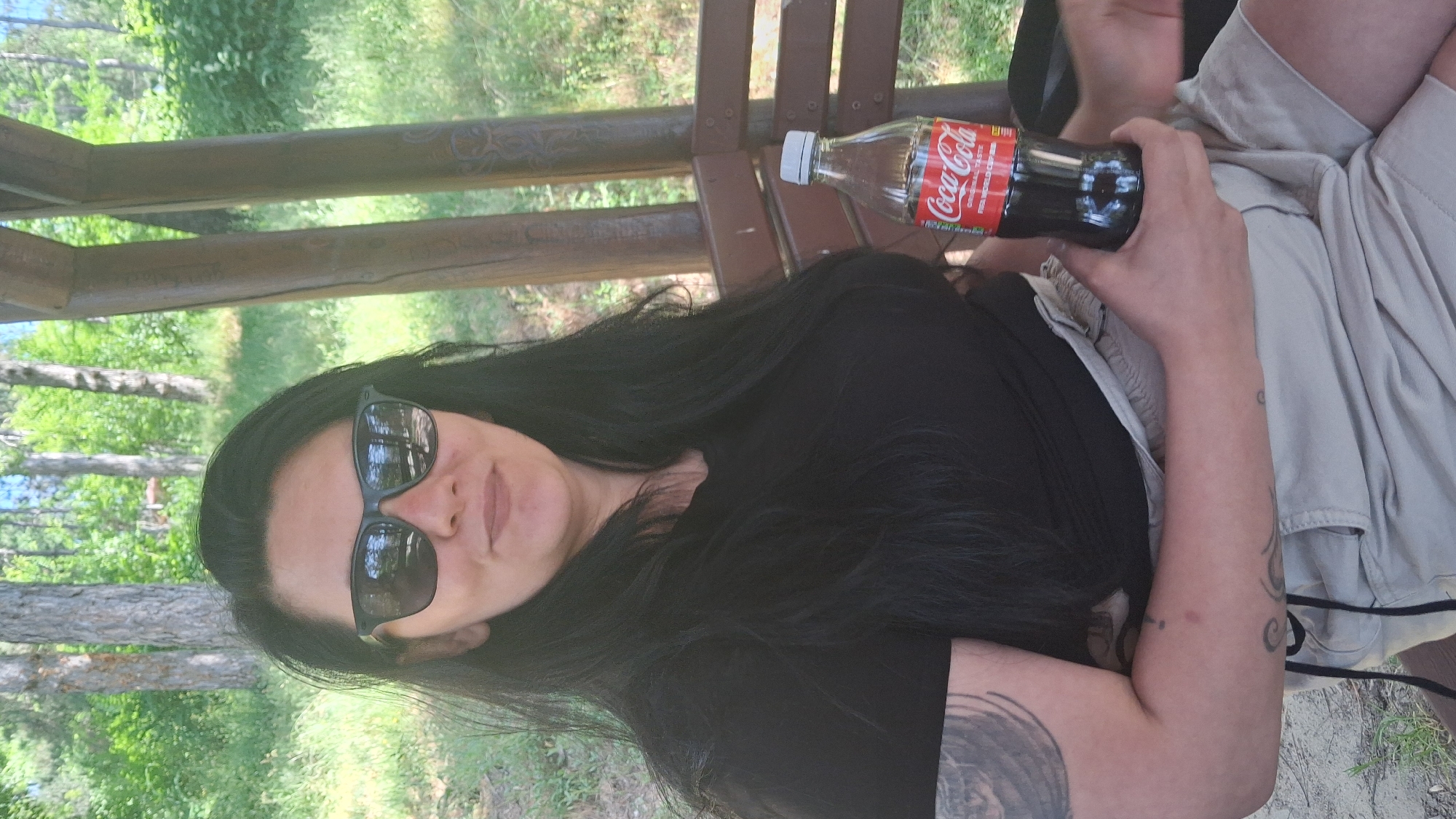 The image shows a woman seated on a wooden bench, possibly in a gazebo or covered structure. She is holding a bottle of Coca-Cola. She has long, dark hair and is wearing sunglasses and a black t-shirt. Her arm has visible tattoos. The background shows a wooded area with trees and foliage. The location appears to be outdoors, possibly in a park or recreational area. The image is probably taken in Gotse Delchev, Bulgaria.