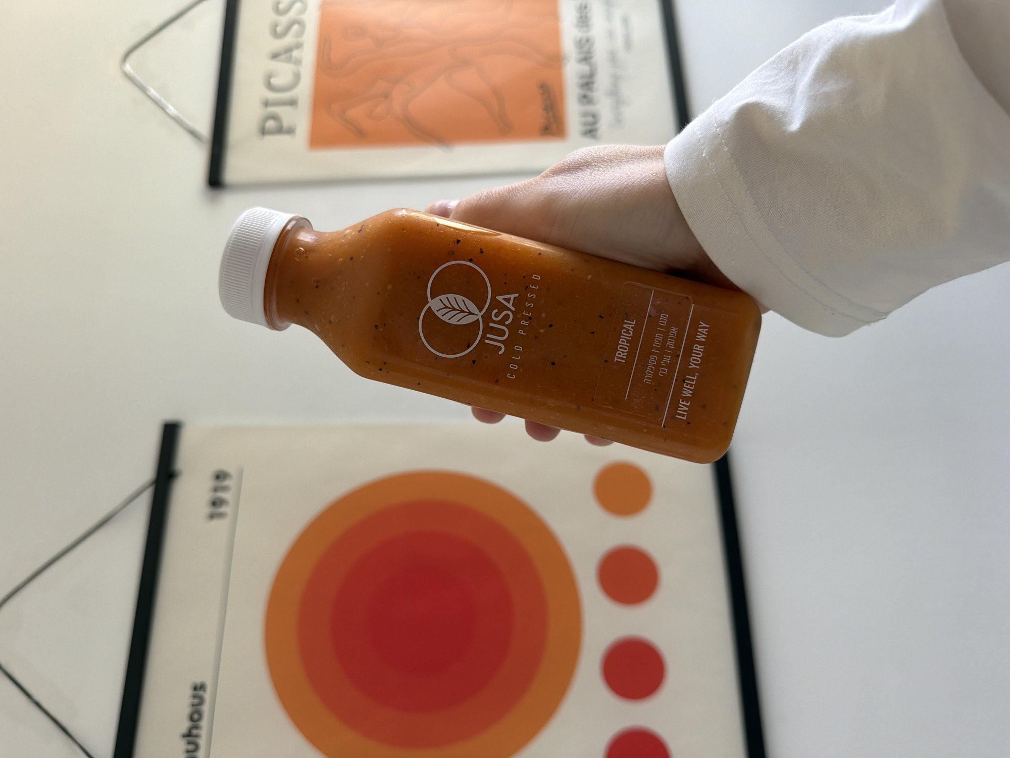 A person's hand in a white sleeve is holding a bottle of "JUSA COLD PRESSED" "TROPICAL" juice. The background contains two posters on a white wall. The first poster reads "PICASS" and "AU PALAIS des." The second poster reads "bauhaus" and "1919" and contains orange circle designs. The bottle label text is written in English and Hebrew. This juice bottle is likely being photographed in Tel Aviv, Israel, based on the Hebrew text on the bottle.