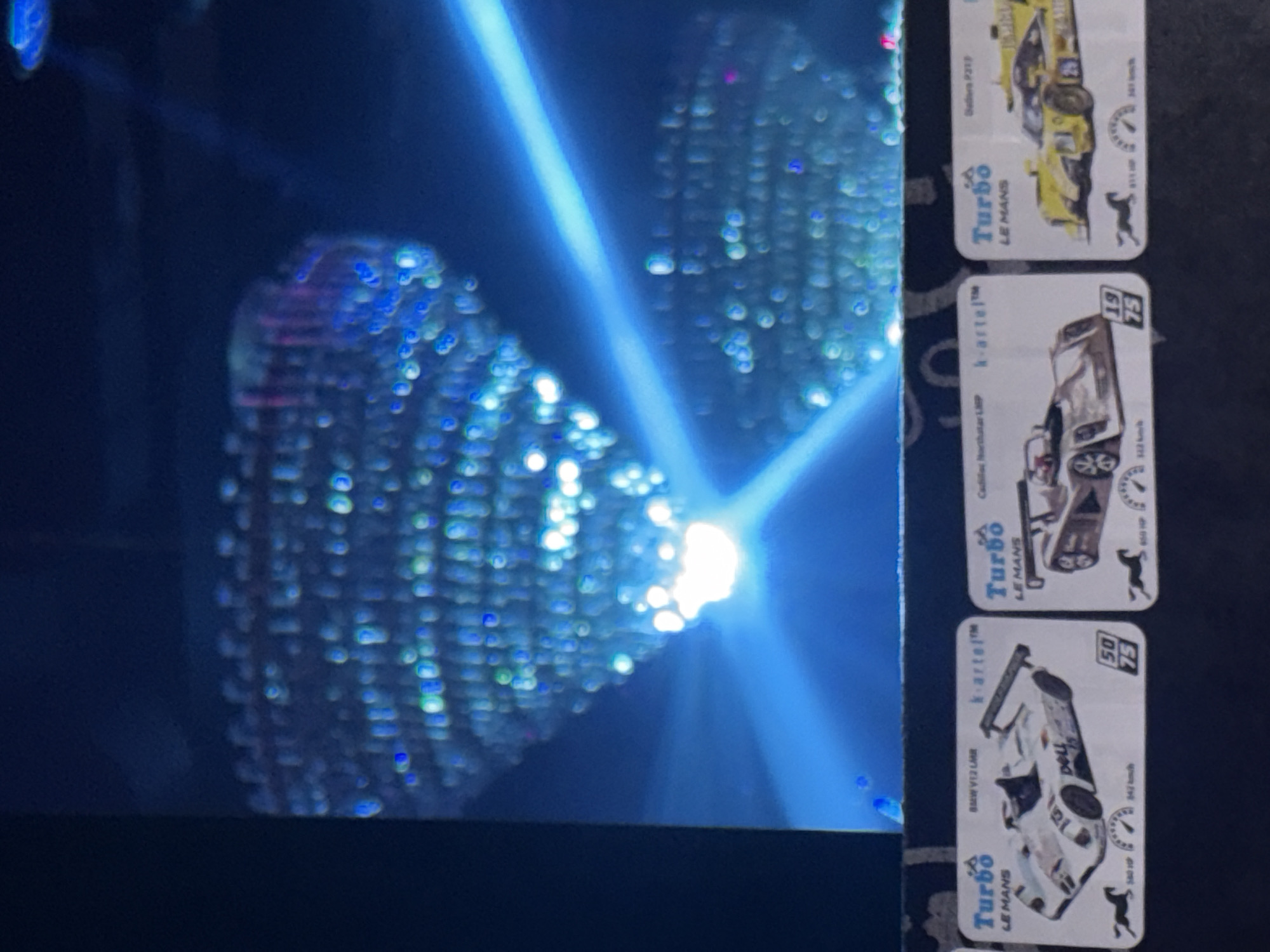 The scene depicts a dimly lit interior environment, characterized by two large, blurred overhead light fixtures resembling chandeliers, emitting blue and white light, and crossed by a prominent blue light beam. In the foreground, three collectible "Turbo LE MANS" racing car cards, produced by "k-artel™", are displayed on a dark, horizontal surface. The leftmost card features a white BMW V12 LMR with "Dallara" branding, indicating 580 HP, 342 km/h, and a "50/75" ratio. The central card shows a brown Cadillac Northstar LMP, listing 650 HP, 322 km/h, and a "19/75" ratio. The rightmost card presents a yellow Dallara P217 with a "25" marking, displaying 615 HP and 342 km/h. Each card includes a horse emblem. No people are visible. The presence of "k-artel™" branded items, a company with a significant presence in Russia, suggests this scene is located within an entertainment facility, arcade, or retail store in Moscow, Russia.