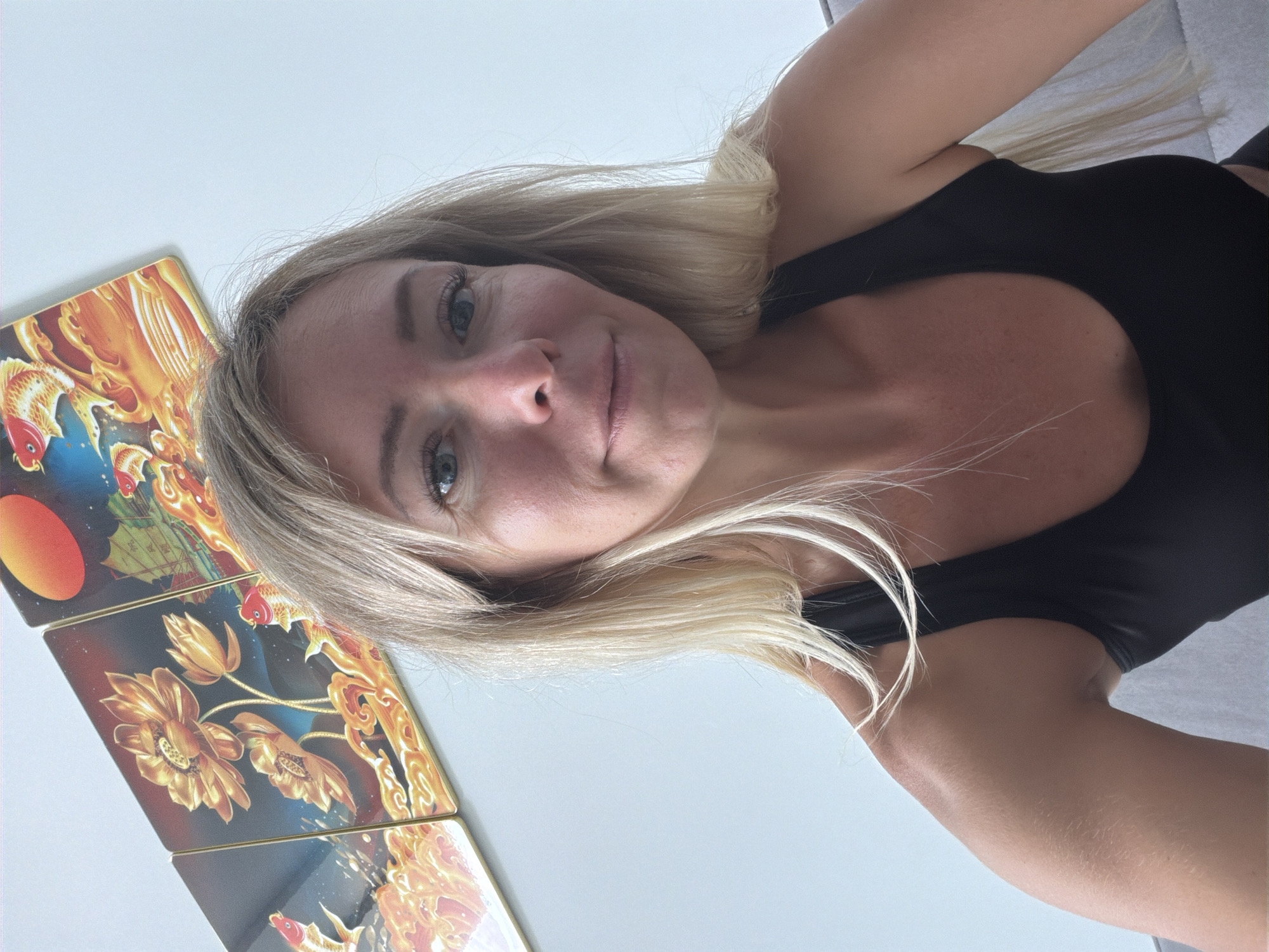 In this close-up selfie, a woman with blonde hair and blue eyes is looking at the camera with a slight smile. She is wearing a black tank top. Behind her, on the wall, is a triptych artwork depicting golden lotus flowers and koi fish against a dark background with a red sun. The scene appears to be indoors, possibly in a home or hotel room. The background wall is a plain light color, and a piece of furniture, possibly a couch, is visible on the right side of the image. The lighting is bright, suggesting daylight. There is no indication of the specific location in Xuân Trường - Đà Lạt, Vietnam, other than the general context of being in a city.