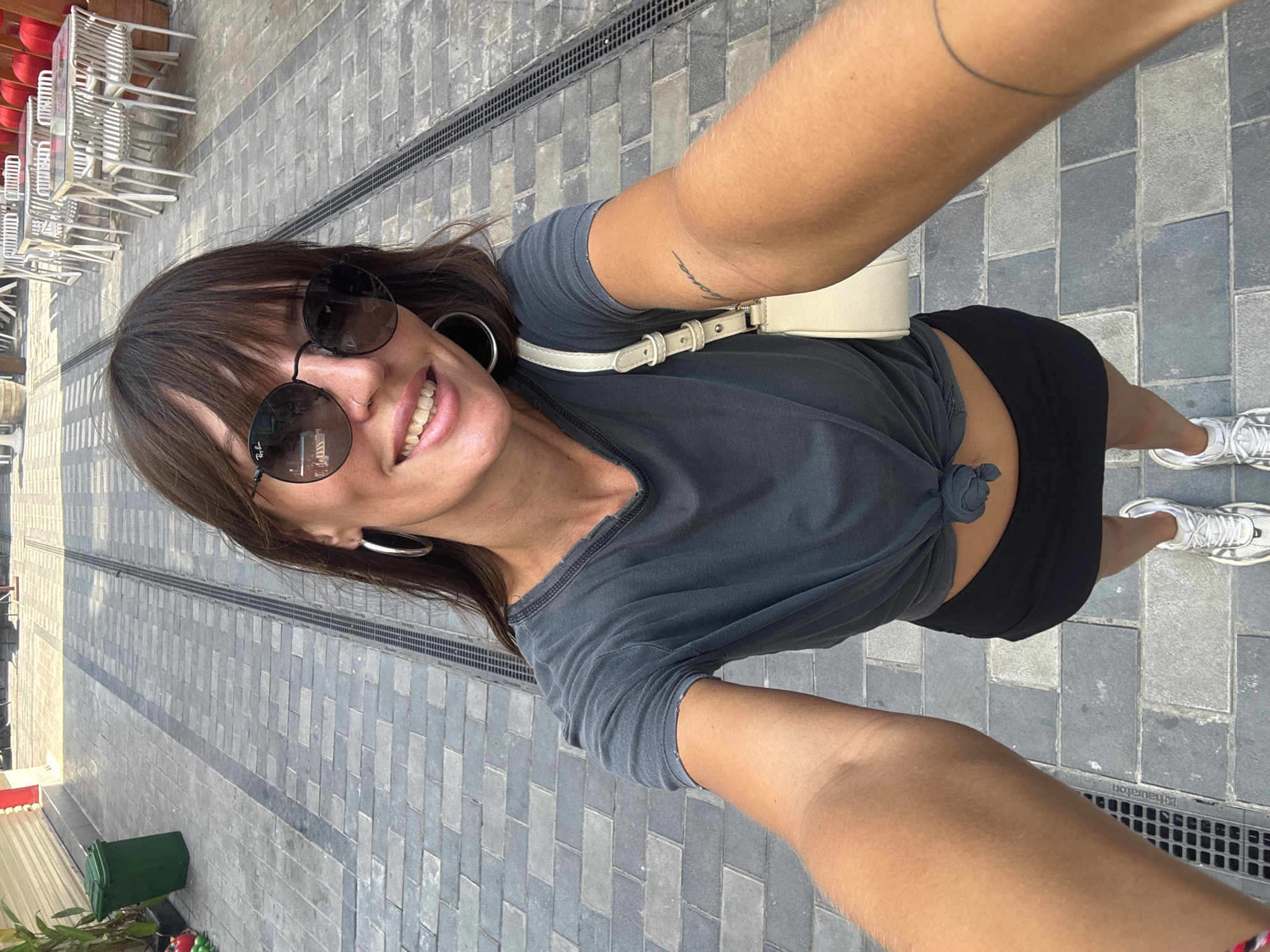 A female subject is captured in a close-up selfie, positioned in an outdoor pedestrian zone within the city of Đà Nẵng, Vietnam. The subject, possessing medium-length brown hair with bangs, is wearing dark, round sunglasses with a visible "Ray-Ban" logo on the left lens. She has a nose piercing, specifically a silver ring in her right nostril, and large silver hoop earrings. Her expression is a smile, revealing her teeth. She is dressed in a dark grey, short-sleeved t-shirt with a distressed neckline, tied at the front to expose her midriff. She wears a black mini-skirt and light-colored athletic shoes with grey accents. A light-colored shoulder bag is draped over her right shoulder. A cursive tattoo is visible on her right forearm, and a thin, partial circular line tattoo is present on her left forearm. Her arms are extended, holding the camera for the selfie.
The environment features a paved ground surface constructed from alternating grey and darker grey rectangular blocks. Two parallel metal drainage grates run longitudinally along the pavement. To the left of the subject, a dark green lidded trash bin is visible, partially obscured by green foliage. To the right, an outdoor dining area is arranged with multiple square tables topped in white and white chairs featuring slatted backs and legs. Further in the background on the right, several red decorative lanterns are visible against a light-colored structure. The scene is brightly illuminated by direct sunlight, casting distinct shadows. No other individuals are discernible in the immediate vicinity.