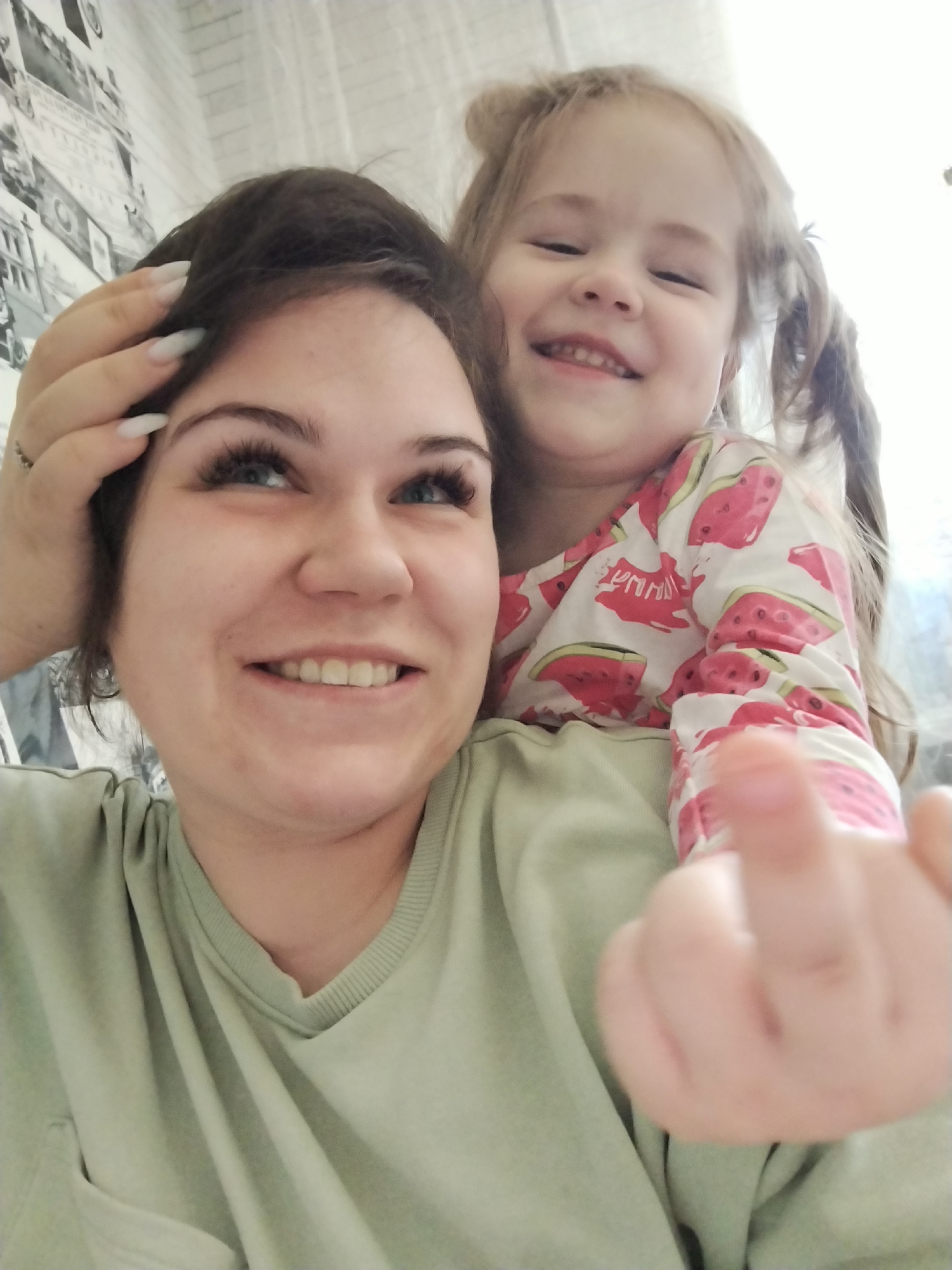 A smiling woman with short dark hair and long artificial nails has her right hand on her head. She is wearing a light green t-shirt. A young girl with blonde pigtails sits on her shoulder, wearing a white and pink watermelon-patterned shirt and smiling. The girl's left hand is outstretched toward the camera. The background features a wall with black and white pictures. The image was taken in the city of Chaikovsky, Russia.