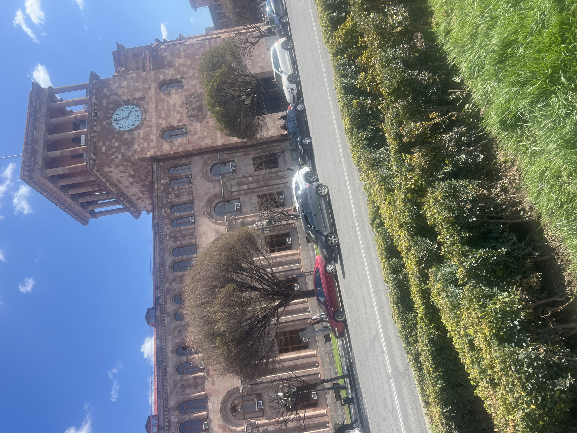 The image shows the building of the Yerevan Municipality in Yerevan, Armenia. The building is made of pink tufa stone and has a clock tower. Several cars are parked along the street next to the building. A row of trimmed bushes separates the street from a grassy area. The sky is blue with some scattered clouds.