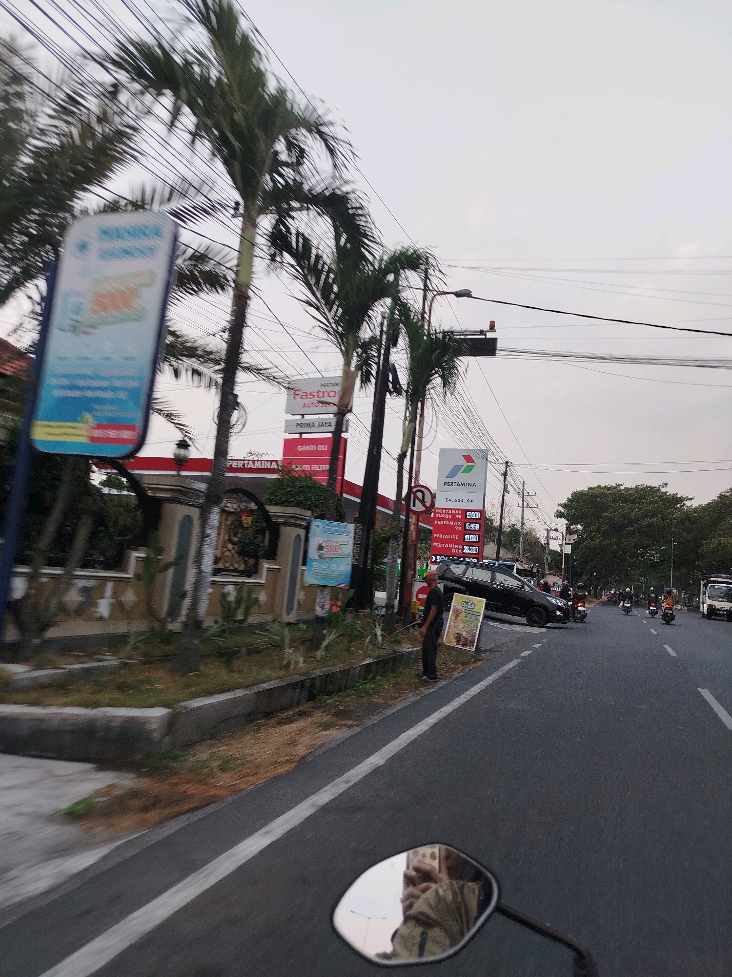 The image depicts a street scene in Cekok, Indonesia, with a focus on a Pertamina gas station and its surroundings. A black car is parked at an odd angle on the side of the road, near a sign displaying prices for different types of gasoline. A man in dark clothing stands near the sign, seemingly observing the scene. Several motorcycles are visible on the road, with some individuals on them. Palm trees line the street, and electrical wires crisscross the sky. In the foreground, a motorcycle's rearview mirror reflects the photographer. The sky is overcast, suggesting it may be late afternoon or early morning. The environment appears to be a typical urban street in Indonesia, with a mix of commercial establishments and traffic.