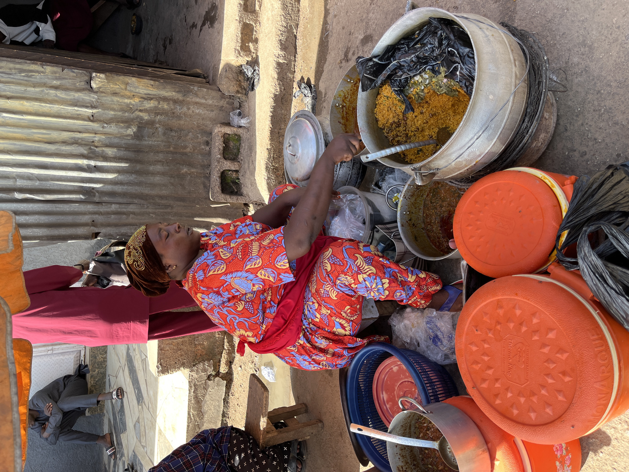 A Black woman wearing a red and blue floral-patterned dress and a brown headscarf is crouched next to a large pot of yellow rice in an outdoor market in Jos, Nigeria. She is scooping food from a pot with a ladle. The market appears to be a busy street scene with other people in the background. The area is dusty and appears to be a typical Nigerian market setting.