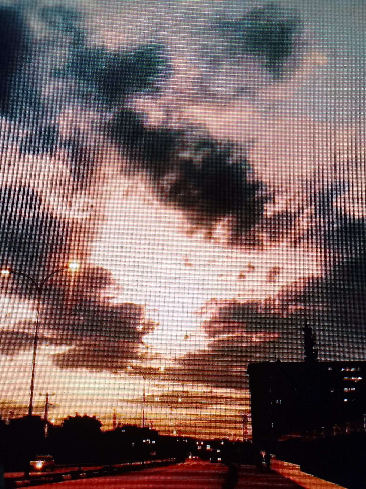 The image depicts an urban landscape at sunset. A road stretches into the distance, flanked by streetlights and silhouettes of buildings and trees. Dark clouds fill the sky, illuminated by the setting sun. The location is a city of unknown. The image is displaying a digital screen.