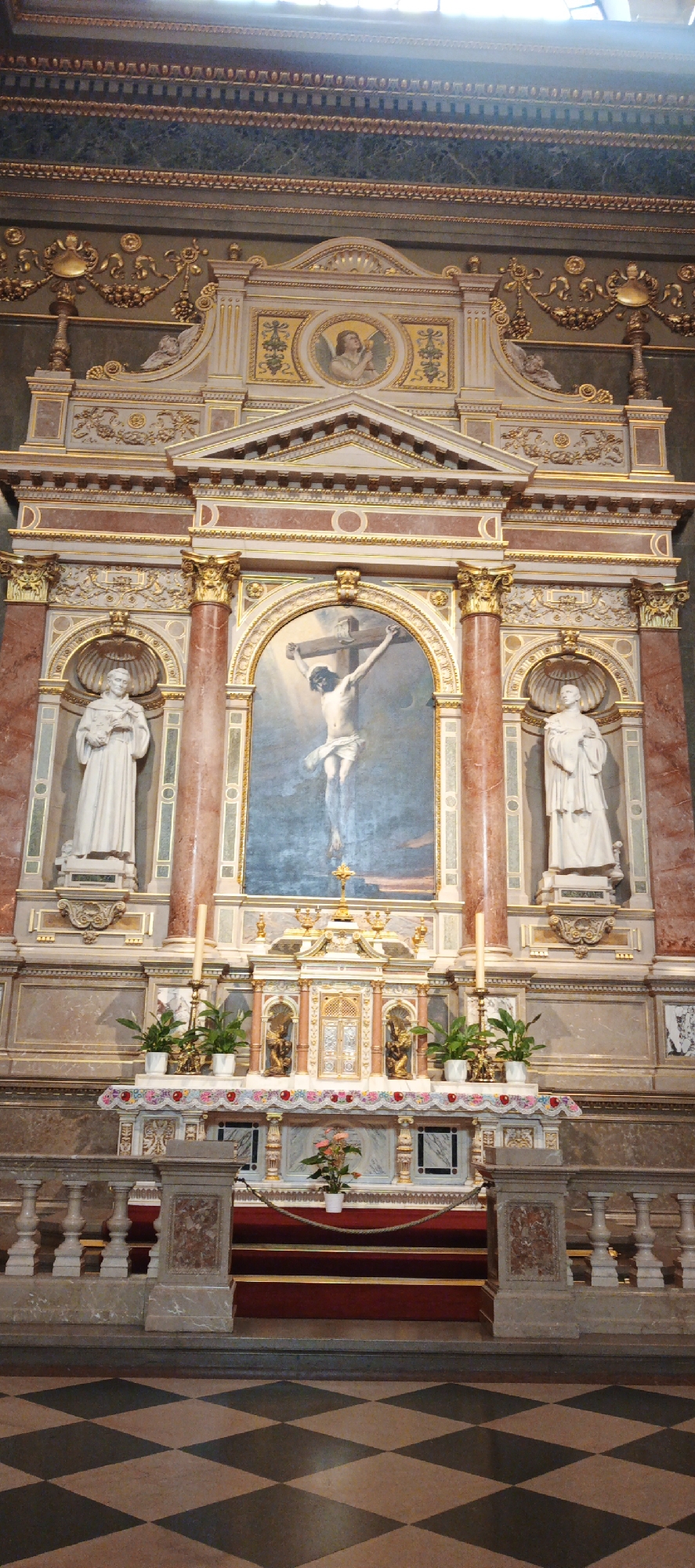 The image shows the main altar of the Inner City Parish Church in Budapest, Hungary. The altar is highly ornate, featuring a painting of the crucifixion of Jesus Christ as the central element. Flanking the painting are two white marble statues of saints, each beneath a small, arched canopy. The altar itself is decorated with a white cloth adorned with colorful floral designs. Potted plants are placed on either side of a small tabernacle on the altar. Two tall candles are positioned towards the front of the altar. The altar is elevated, with a red carpeted step leading up to it. The floor in front of the altar is tiled in a black and brown checkered pattern. The overall style of the altar and its surroundings is Baroque or Rococo, characterized by elaborate ornamentation and gilded details. The light suggests the image was taken during daytime.