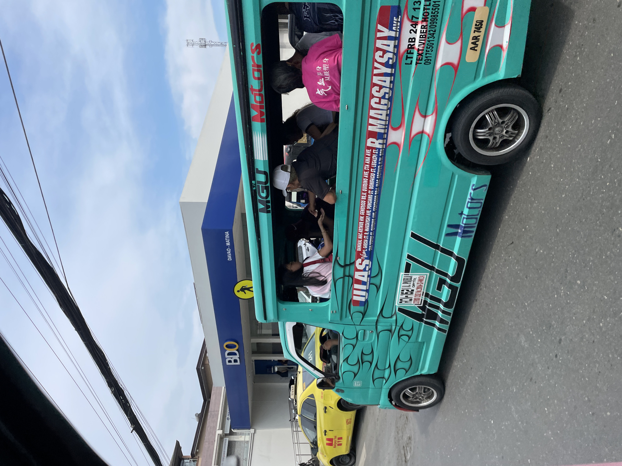 This image captures a daytime street scene in Matina, Davao City, Philippines. The primary focus is a teal-colored multi-cab (a type of public utility vehicle) moving along an asphalt road, with a BDO bank branch visible in the background.
The multi-cab, decorated with white and red flame decals, is labeled with "MGU Motors," "ULAS," "R. MAGSAYSAY AVE.," and several other street names indicative of its route (e.g., "BANGKAL MAC ARTHUR HWY., GENEROSO BRGY. QUIRINO AVE., STA. ANA AVE."). It also displays operational information such as "LTFRB 24/7 13 TEXT/VIBER HOTLI 09175561342/09985501," "ROH-162 I.A. PADILLA OPTR.," "LTFRB CASE NO. 2018-12-00124," and a route or vehicle number "AAR 7450." The vehicle is equipped with chrome-finished multi-spoke wheels.
Inside the open-sided passenger compartment of the multi-cab, multiple individuals are seated. A woman with long dark hair, wearing a light-colored top and a dark strap, is visible near the front, looking forward. A man with a white cap and a dark t-shirt is seated, interacting with someone towards the center. Another passenger, with gray hair, wears a pink t-shirt with Chinese characters on the back (气血养身 从根塑身), also seated and looking forward. At least two other passengers are partially visible, indicating a full vehicle.
Behind the multi-cab, a yellow sedan taxi, branded "Maligaya Taxi" and numbered "1673," is partially visible on the road.
The BDO bank branch in the background is a blue and white building with the "BDO" logo prominently displayed. Text below the logo reads "DAVAO - MATINA," confirming the specific location within Davao City. A yellow pedestrian symbol is affixed to the bank's exterior wall. Automated Teller Machines (ATMs) are visible through the glass entrance.
Overhead, a dense bundle of black utility wires and thinner individual lines crisscross against a partially cloudy blue sky. To the far right in the background, a metal lattice communication tower is visible. The scene suggests a typical urban environment with active transportation and commercial activity.
