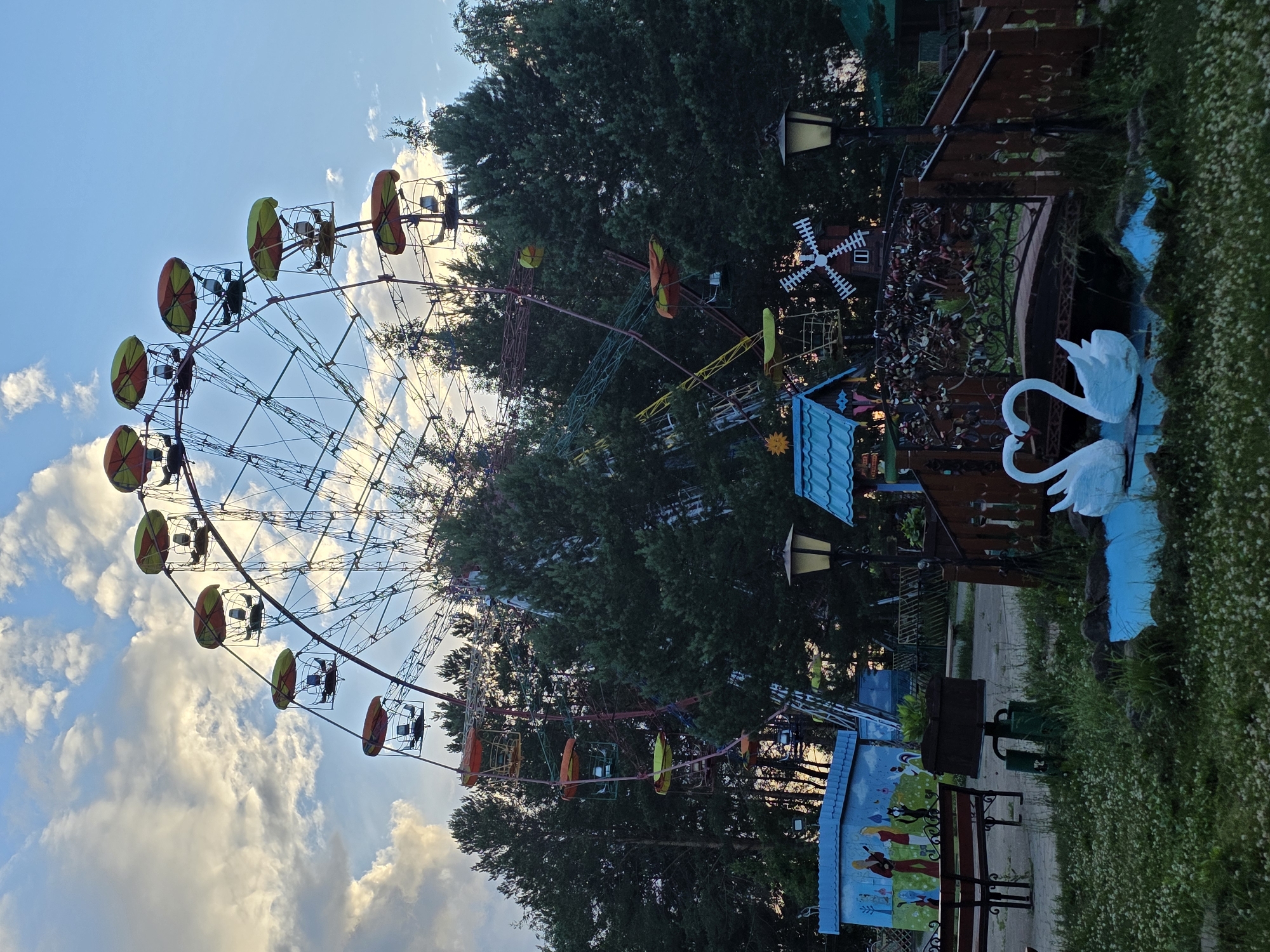 The image depicts a Ferris wheel at an amusement park in Chaikovsky, Russia. The Ferris wheel has yellow and red gondolas, some of which appear to be occupied by people. The Ferris wheel is set against a backdrop of green trees and a blue sky with scattered clouds, suggesting it is daytime.
In the foreground, to the right of the Ferris wheel, there is a pond with two white swan sculptures. There are also decorative elements like a miniature windmill and lampposts. A colorful mural depicting people and animals is visible on a structure below the Ferris wheel, and a park bench is placed in front of it. The overall impression is of a cheerful and lively amusement park environment. The time of day appears to be late afternoon or early evening due to the lighting and the colors of the sky. The weather seems pleasant, with a mix of sun and clouds.
