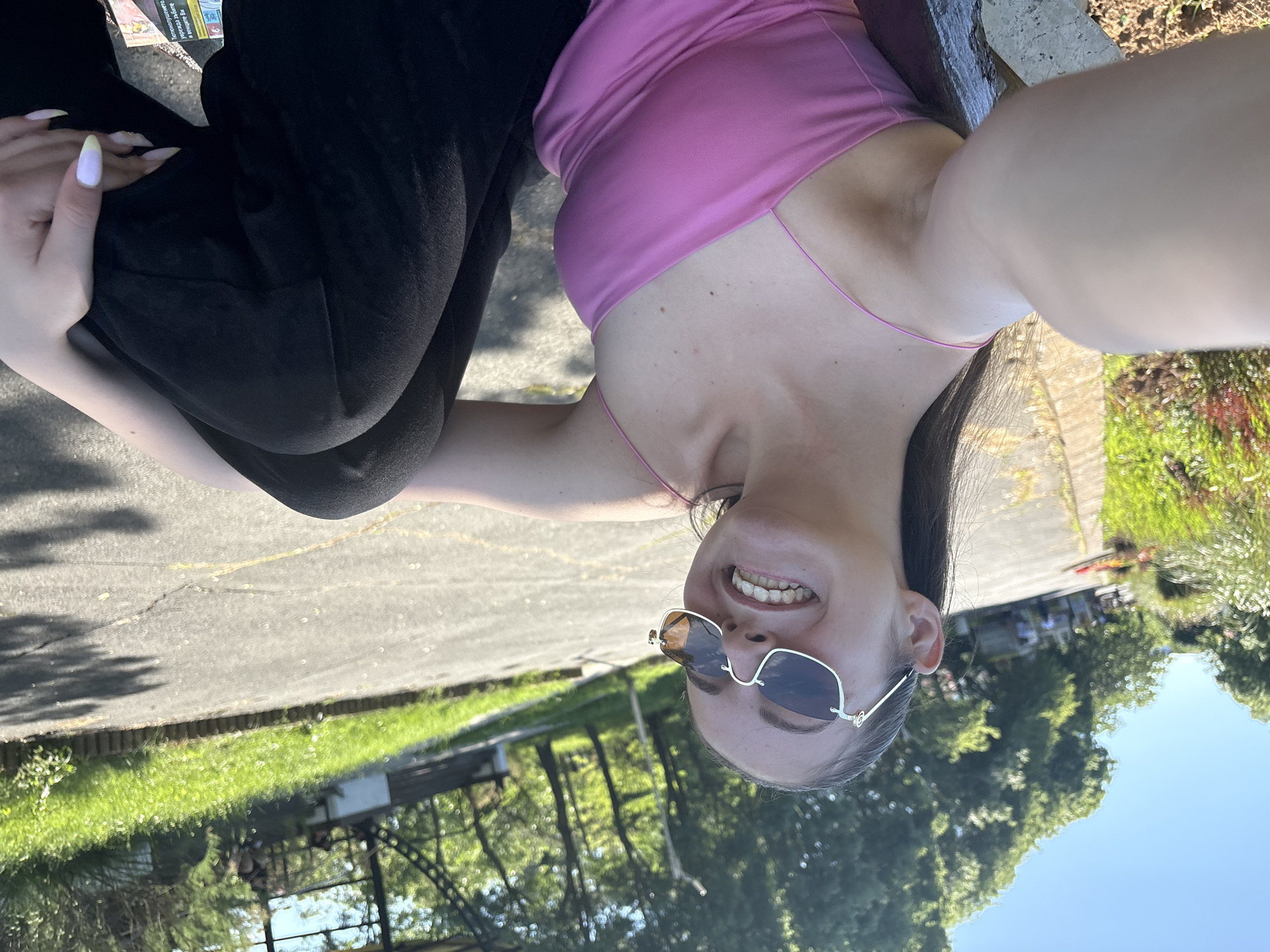 A young woman in a pink top and black pants is taking a selfie outdoors. She is smiling and wearing sunglasses. The background shows a park-like setting with trees, grass, a paved area, and a fence. People are visible in the distance. The image is rotated, and the location is likely in Tsarevo, Bulgaria.