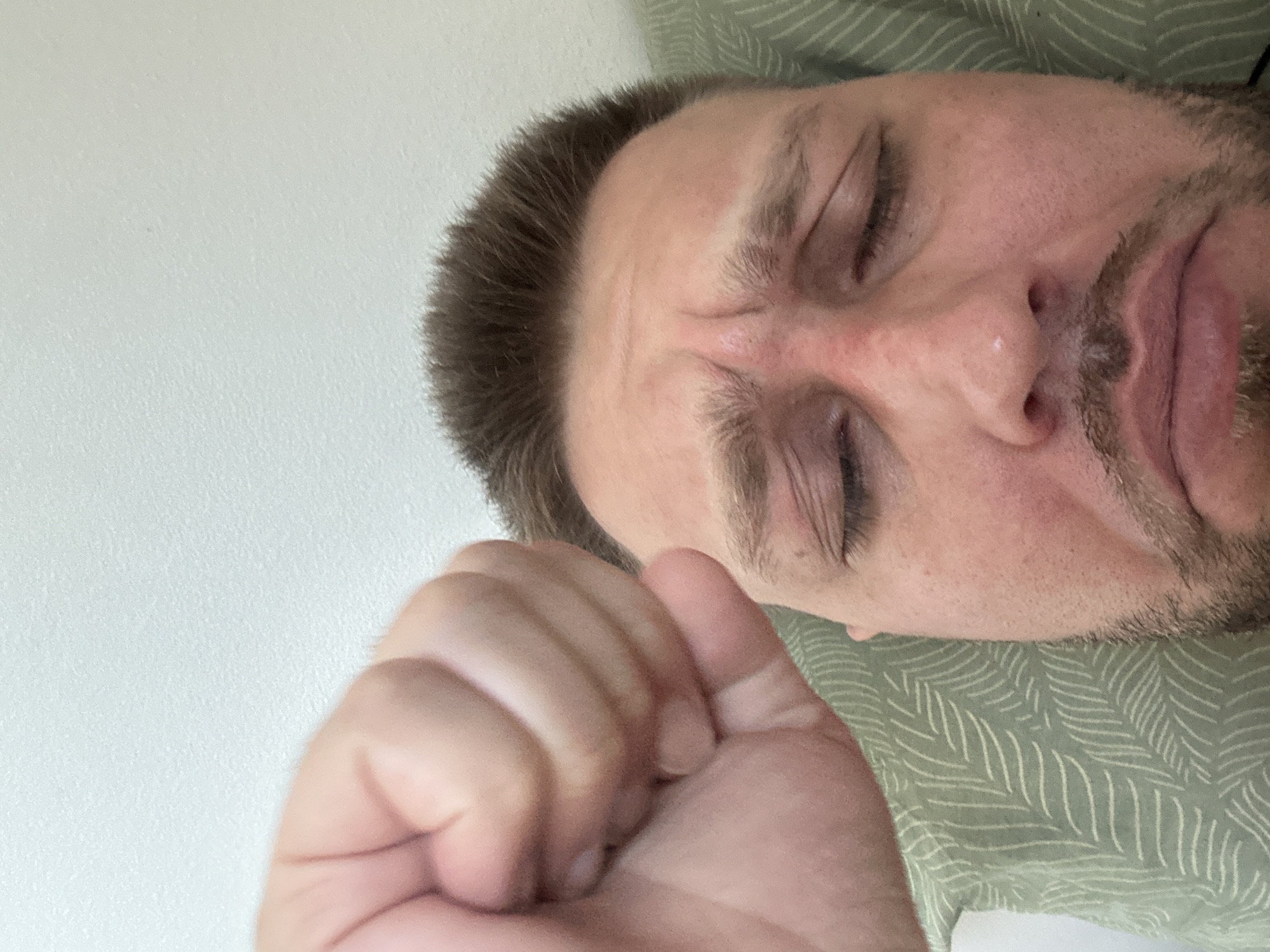 A close-up, high-angle shot shows a man's face, eyes closed, with his forehead creased in distress or pain. His right fist is clenched and held close to his face, obscuring part of his forehead and hair. He is lying on a light green pillow with a white leaf-like pattern. The background is a textured, off-white wall. The man has short brown hair and stubble on his chin. The lighting is even and appears to be from an overhead source. There is no indication of the man's location within Zvezdara Urban Municipality, Serbia.