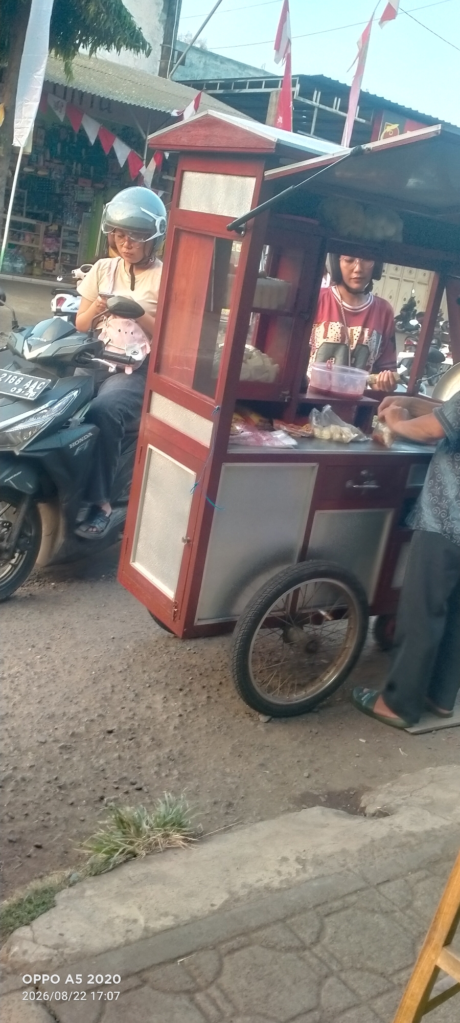 The image depicts an outdoor scene in Kedung, Indonesia, likely a street or marketplace, with a food cart as the central focus. A woman wearing a helmet and glasses is seated on a motorcycle, looking down at her phone. She has a pink plush toy attached to her backpack. Next to the motorcycle, a wooden food cart with red and silver accents is open, displaying various food items in plastic bags and containers. Another person, partially visible, is working behind the cart, handling food. The cart has a wheel on the right side. In the background, buildings and trees are visible, along with red and white triangular flags, possibly indicating a celebration or national holiday. The ground is a mix of dirt and paving stones. The timestamp on the image reads "OPPO A5 2020 2026/08/22 17:07".
