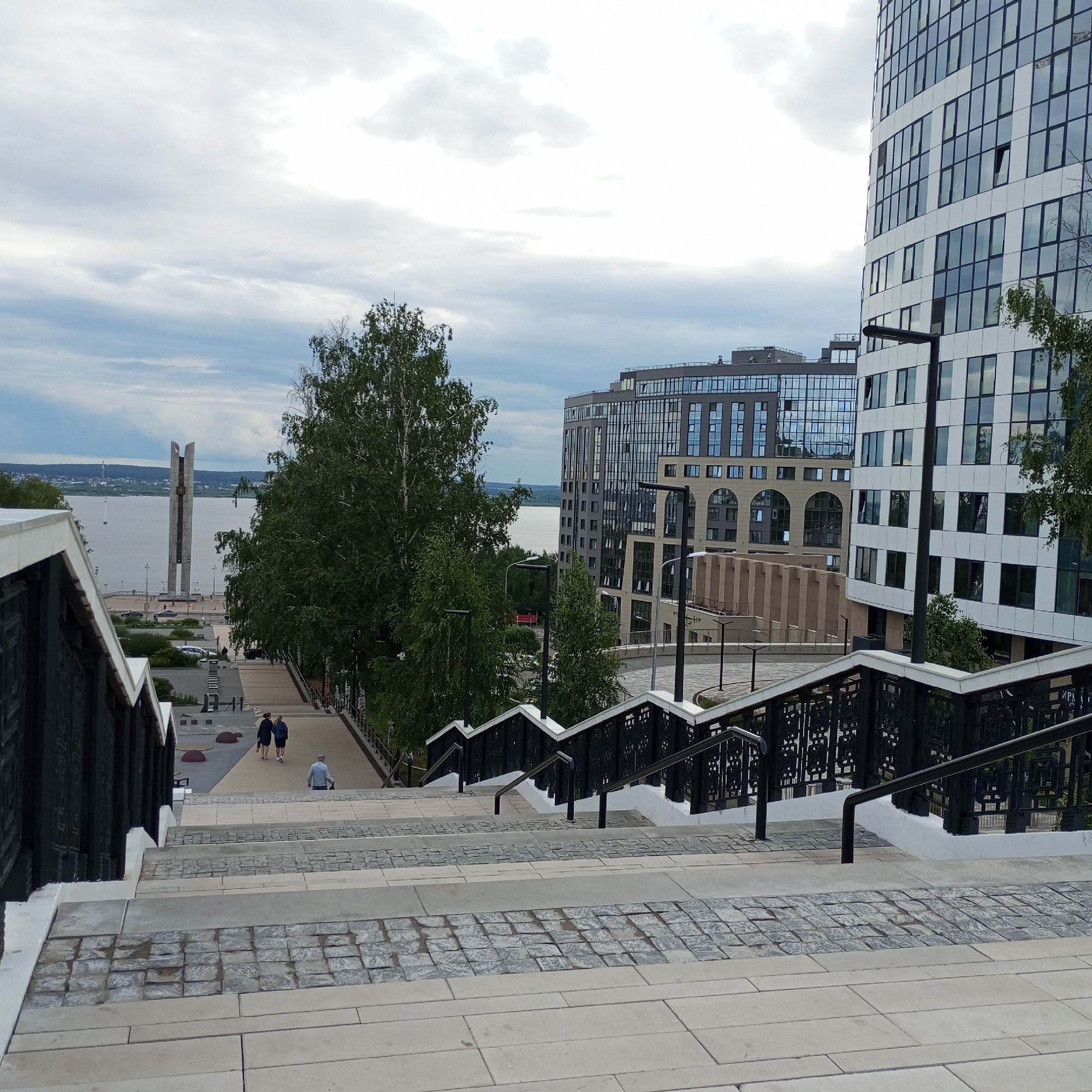 This detailed image depicts a modern urban embankment area in Izhevsk, Russia, specifically overlooking the Izhevsk Pond/Reservoir with the "Friendship of Peoples Monument" visible in the distance.
In the foreground, a wide, multi-tiered staircase descends towards the water. The stairs are constructed from light-colored concrete or stone slabs, with alternating bands of grey cobblestone pavers. Both sides of the staircase feature black metal railings with geometric patterns, supported by white concrete bases and equipped with simple black handrails.
Further down the slope, on a wide paved promenade, two individuals in dark clothing are walking away from the viewer towards the monument. To their left, a single individual wearing a light blue shirt and dark pants is seated on a low wall or step, facing the direction of the water. Green-leafed trees, appearing to be birch, line the sides of the promenade and the steps.
In the midground, the expansive Izhevsk Pond/Reservoir stretches horizontally. A single white sailboat is visible on the water towards the left. On the far side of the pond, distant cityscapes and a low-lying treeline define the horizon under an overcast sky filled with light grey and white clouds. The prominent "Friendship of Peoples Monument" (also known as the "Monument of Eternal Glory"), a tall, slender, V-shaped or split-pillar structure, stands on the embankment directly across the water from the viewer's current position, featuring what appears to be a clock face on its visible side. Cars and streetlights are visible on the road and promenade surrounding the monument.
On the right side of the image, two large, modern buildings dominate the urban landscape. The building closer to the foreground is a tall, curved structure with a facade composed of white panels and numerous rectangular windows, some of which are ajar. A modern black streetlight is positioned in front of this building. Behind it and slightly to the left, another multi-story modern building features a grey facade with a grid pattern of windows, including several distinct arched windows on its lower levels. More trees are interspersed between these buildings.