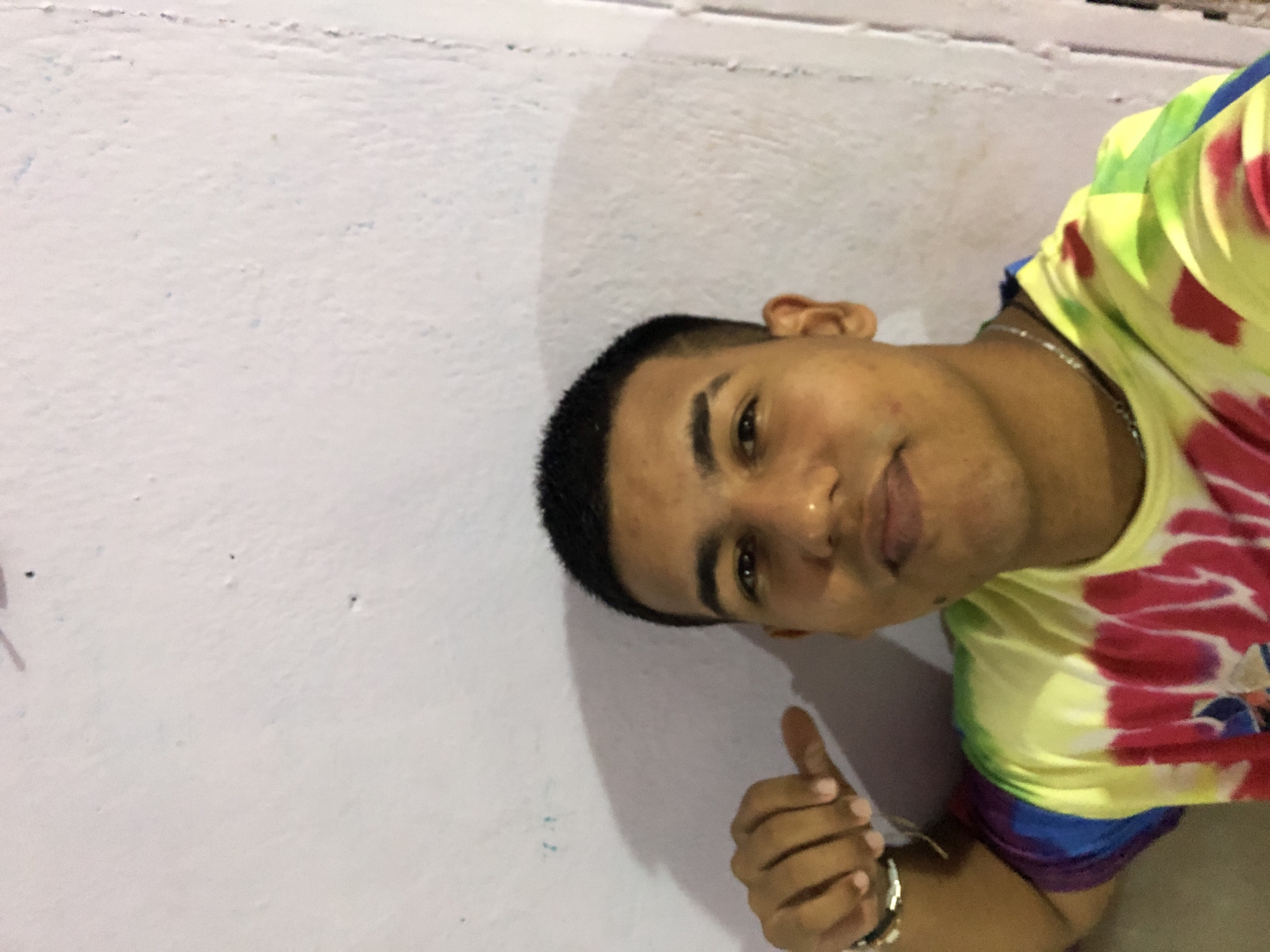 A young man poses indoors with his thumb up. He has short black hair and is wearing a colorful tie-dye shirt and a silver bracelet. A silver necklace is visible around his neck. The background is a plain, light-colored wall. The city of Altagracia, Venezuela is not discernible from the image.