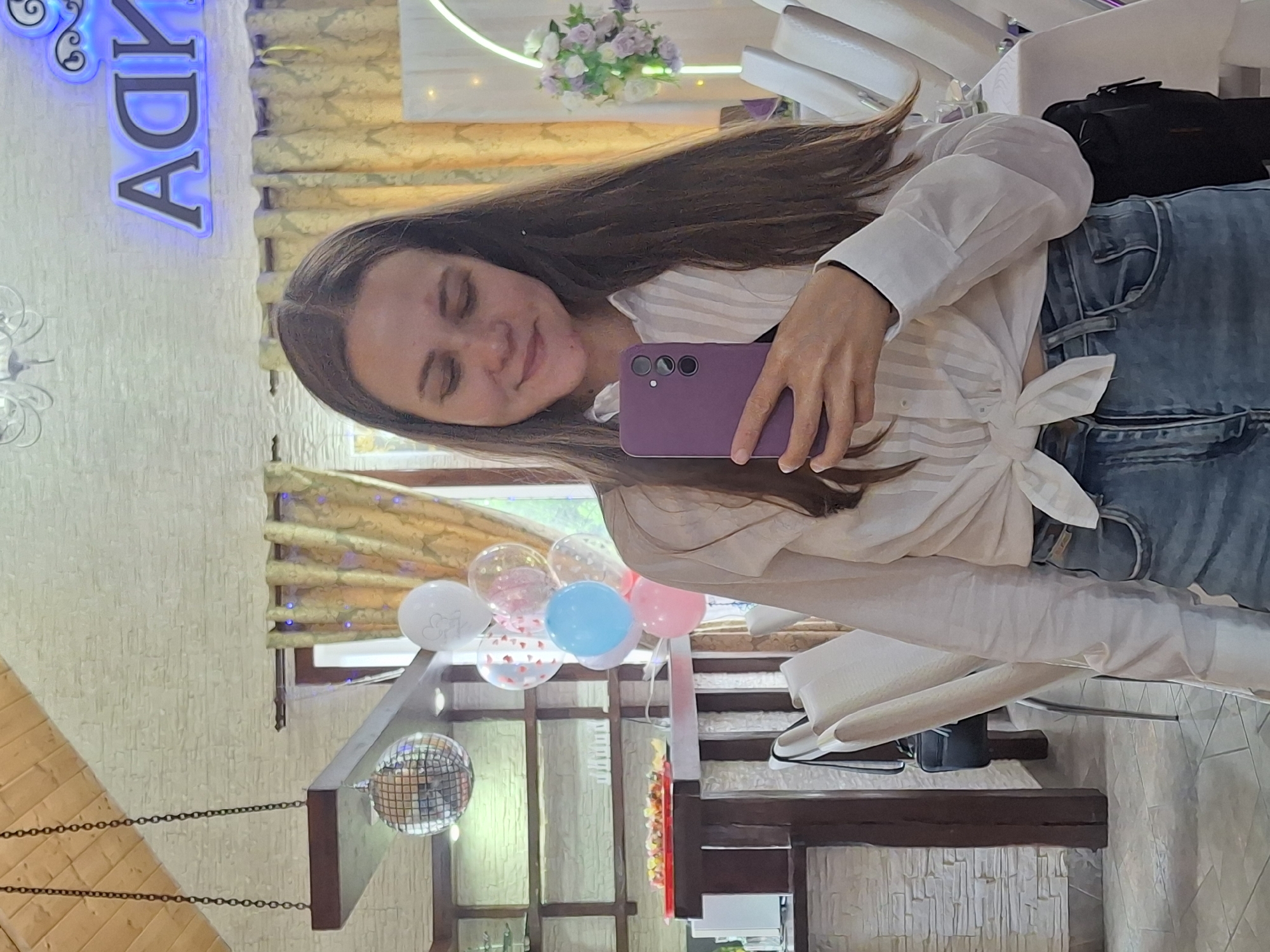 A young woman with long, brown hair is taking a selfie in a dimly lit, indoor venue. She is wearing a white, knotted shirt and blue jeans, and holding a purple phone. In the background, there are balloons, a disco ball, and a neon sign that reads "АДМИРАЛ". This suggests the location is likely the Admiralsky restaurant in Chaikovsky, Russia, a place known for its festive atmosphere and capacity to host events. The overall impression is of a casual gathering or celebration.