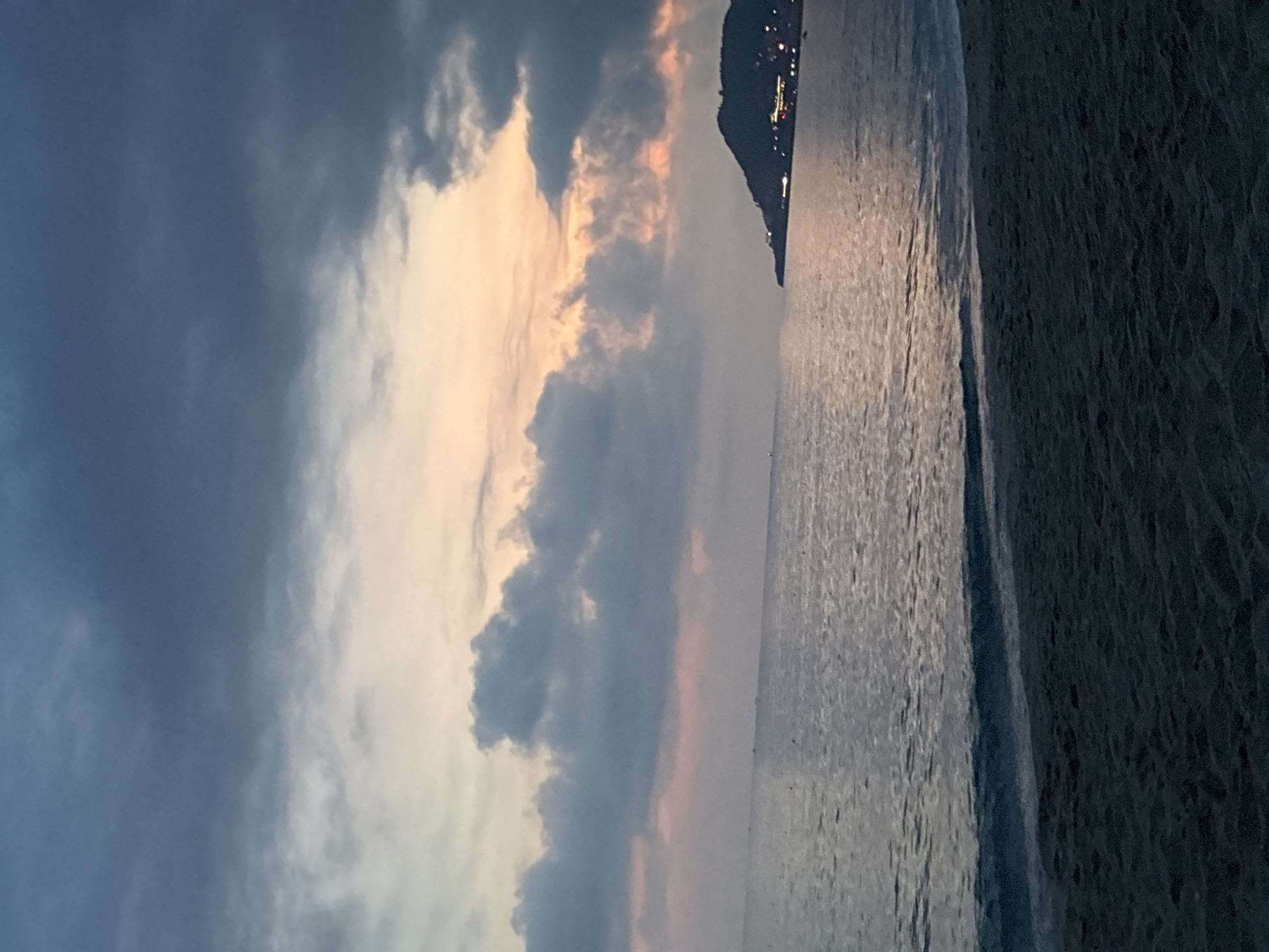 The image captures a serene evening view of a beach in Alanya, Turkey. The foreground features dark, wet sand, indicative of the tide receding. A gentle wave breaks on the shore, its white foam contrasting with the darker sand and the tranquil sea beyond. The sea itself is calm, reflecting the muted colors of the twilight sky. In the distance, a dark, hilly peninsula juts out into the water, its slopes dotted with lights from buildings, suggesting a town or resort area. The sky above is a dramatic canvas of clouds, with patches of soft pink and orange hues from the setting sun peeking through the darker, brooding grey clouds. The overall atmosphere is peaceful and contemplative, evoking a sense of twilight tranquility.