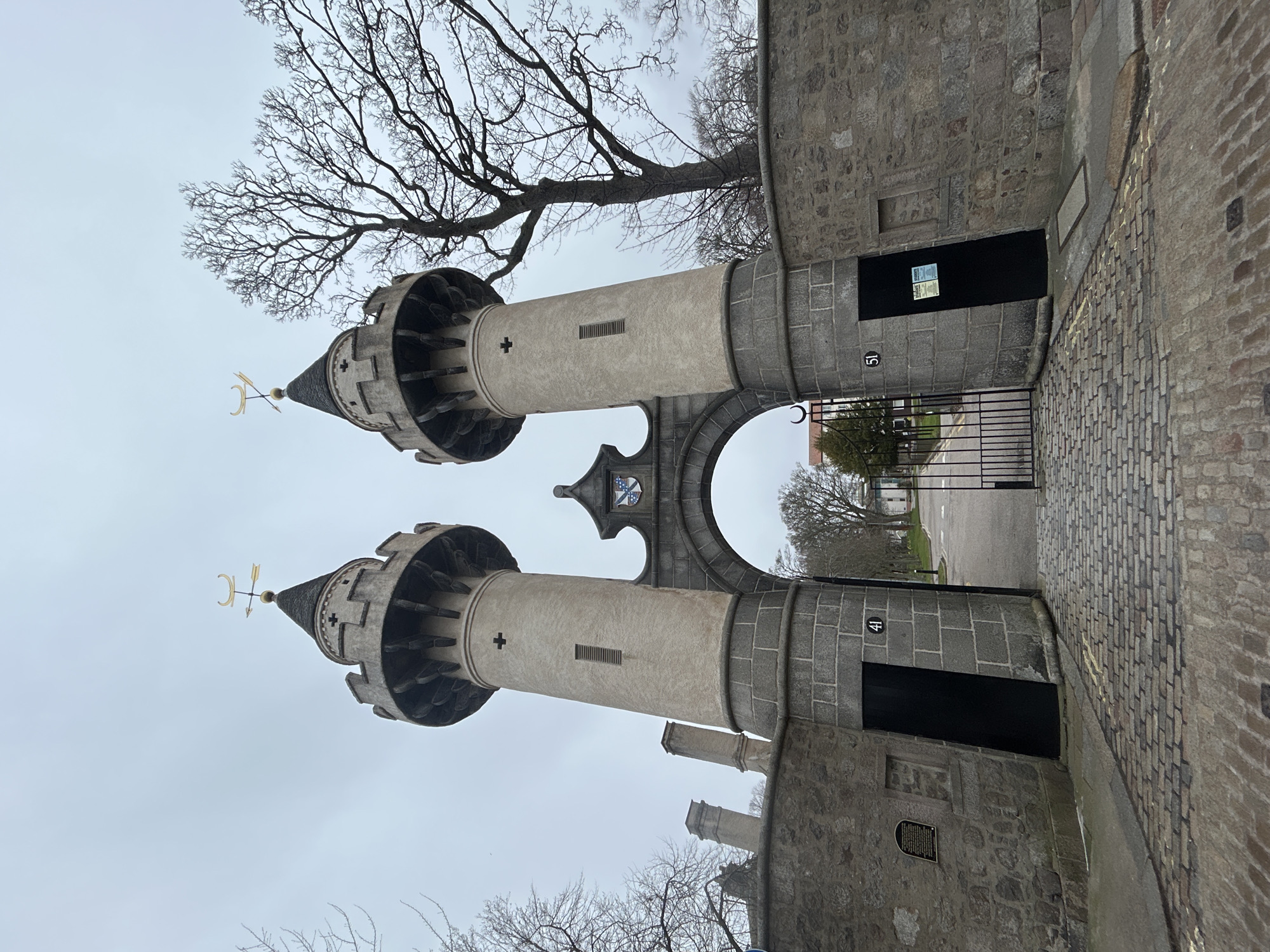 The image shows the Brig o' Balgownie, a medieval bridge located in Aberdeen, Scotland, United Kingdom. The bridge's towers are visible, featuring conical spires topped with weathervanes. The towers are constructed of gray stone and are adorned with decorative elements. An arched opening in the center allows passage through the structure, revealing a view of a street with a barrier. The area surrounding the bridge is paved with cobblestones. A bare tree branch appears in the upper portion of the image, and the sky is overcast. Number plaques "41" and "51" are seen.