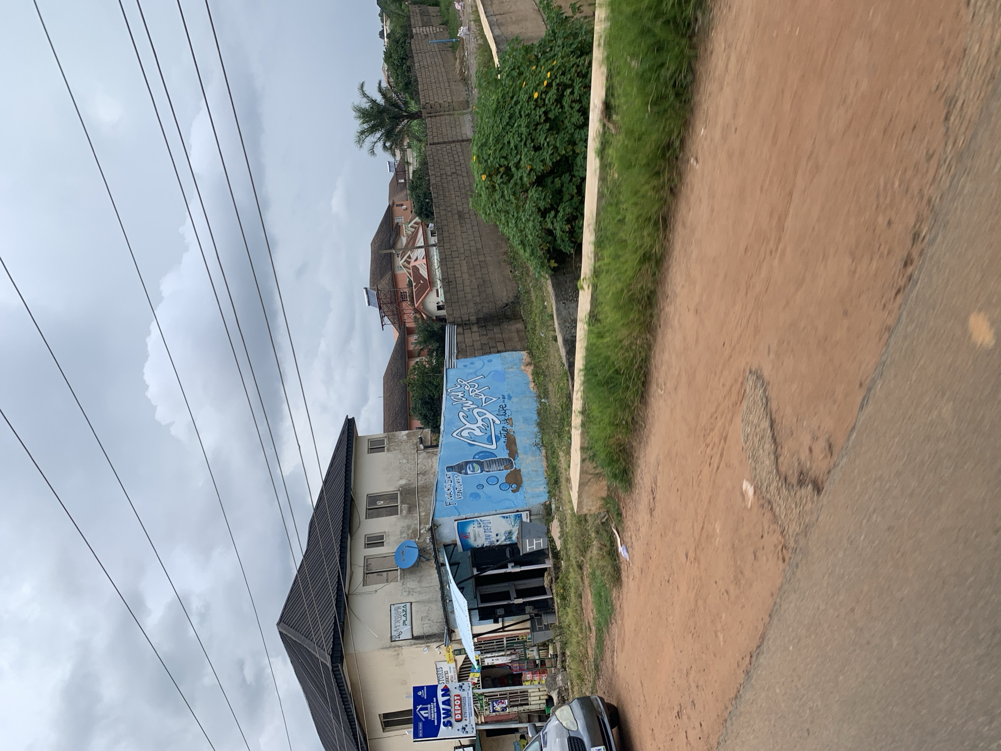 The image captures a street scene in Gura Top, Nigeria, under an overcast sky. In the foreground, a dirt road with some asphalt patching is visible. To the left, the front of a gray car is partially visible.
The main focus of the image is a two-story building with a dark, sloped roof. A blue satellite dish is mounted on the side of this building. Adjacent to the building is a small, blue-painted structure, possibly a kiosk or shop, with a light blue awning. This structure is adorned with graffiti depicting a bottle and the text "SWAN DEPOT," along with other writing that is partially obscured. A sign for "SWAN STORES COSMETICS" is also visible on the left side of the building.
Beyond the immediate structures, a brick wall separates the property from further buildings and vegetation. These buildings appear to be residential, with some having solar water heaters on their roofs. A palm tree is visible to the right of these buildings. A large, lush green bush with yellow flowers is situated in front of the brick wall. The overall impression is a snapshot of a commercial and residential area in Nigeria.