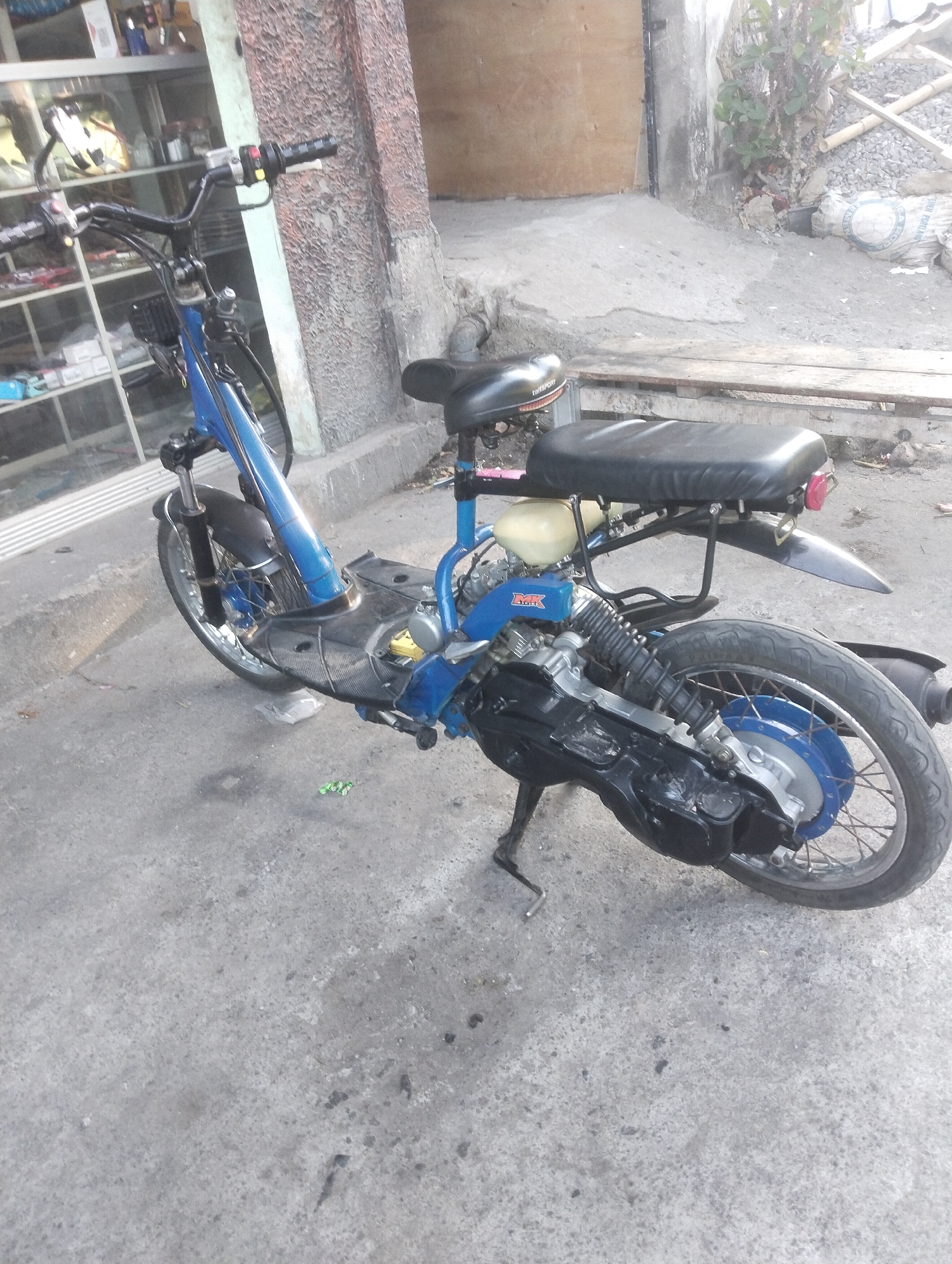A blue and black scooter is parked on a concrete surface in front of a building. The scooter has a black seat, a black engine cover, and a black fender. The building has a textured, light-colored wall and a wooden door. There are some plants growing near the building, and a pile of gravel and bags of cement in the background. The scene appears to be in an outdoor, urban environment.