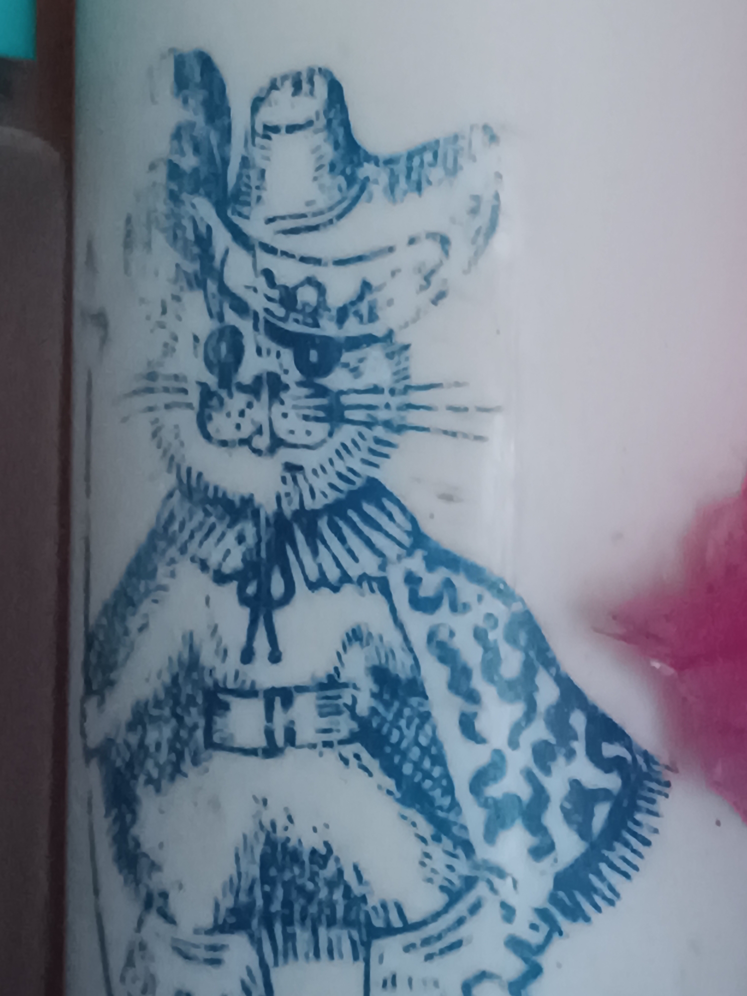 The image shows a close-up of a blue ink drawing on a white surface, possibly a mug or a piece of pottery. The drawing depicts a cat wearing a hat and a cape. The cat has a distinguished appearance, with whiskers and prominent eyes. The hat is wide-brimmed and adorned with a feather. The cape has a patterned design. The drawing style is detailed, with fine lines and shading used to create texture and depth. The background is plain white, with some hints of a pink object in the upper right corner. The overall impression is one of a whimsical and charming illustration. The image does not contain any information about the city of Vypasne, Ukraine.