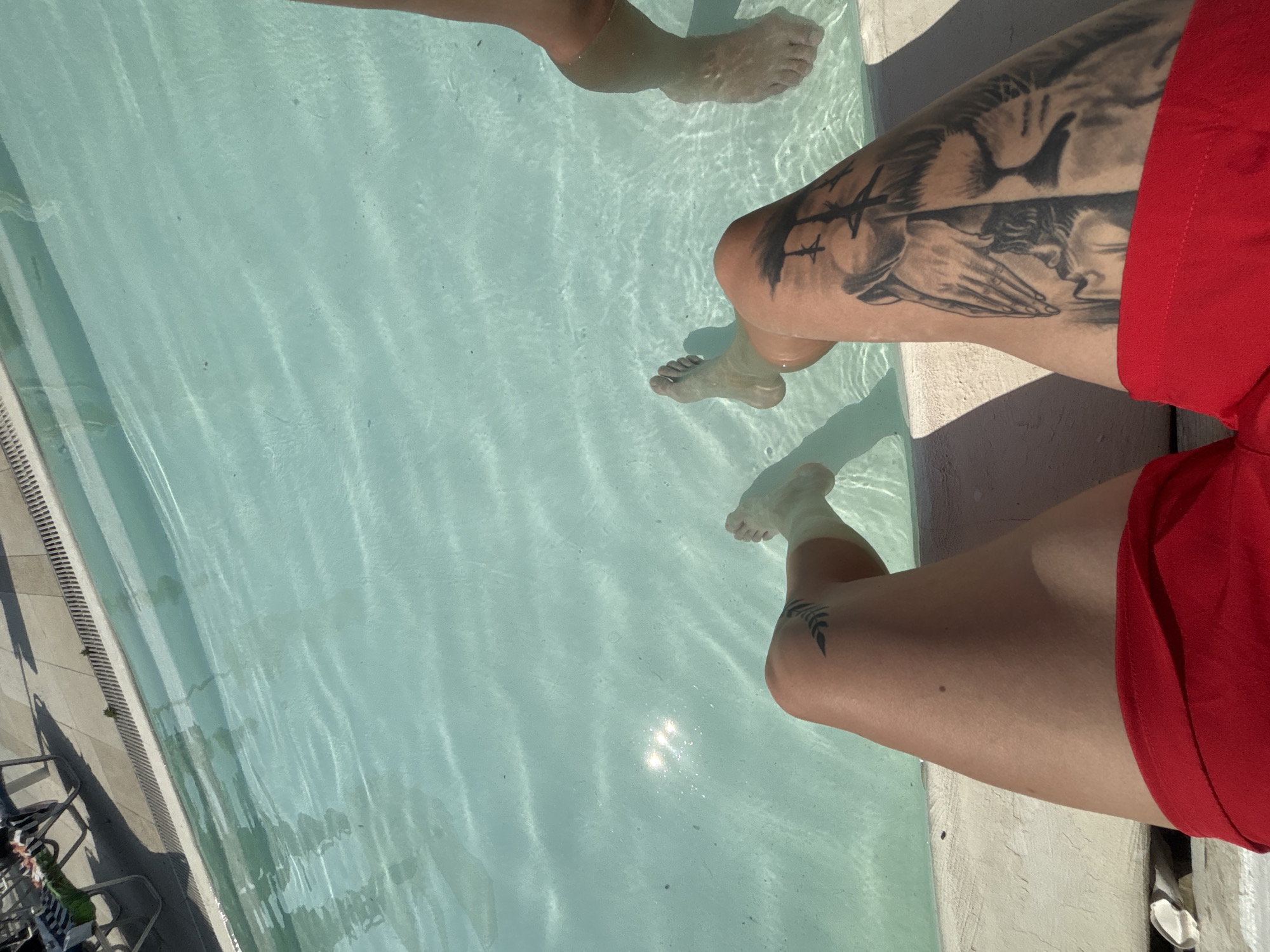 The image shows two pairs of legs partially submerged in clear blue water, likely a swimming pool. The legs are positioned at the edge of the pool, with one person's legs more prominently in the foreground. This person is wearing red swimwear and has tattoos on their left thigh and calf. The other person's legs are visible to the right, also in the water. The surrounding area appears to be a paved poolside with lounge chairs visible in the background, suggesting a recreational setting. The lighting indicates it is daytime. The location is described as Byala, Bulgaria.
