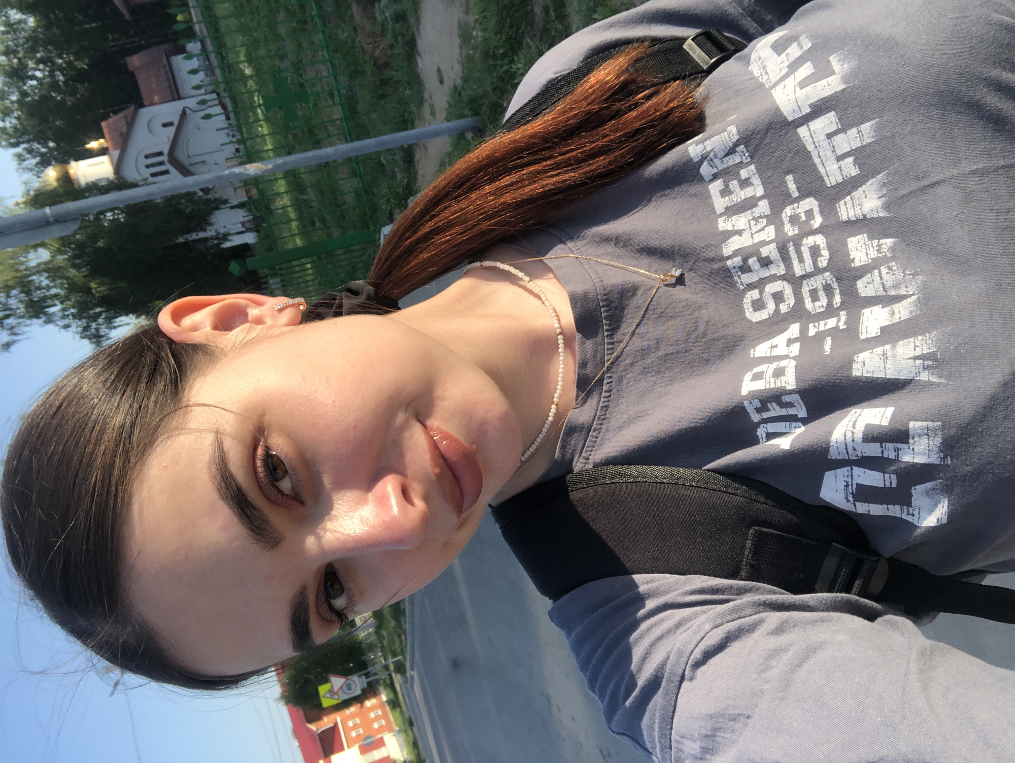 A young woman is taking a selfie outdoors on a sunny day. She has brown hair pulled back, fair skin, and is wearing a grey t-shirt with white text and a black backpack. She is also wearing jewelry: a pearl necklace, a gold pendant necklace, and a bar-shaped earring in her upper ear. The background appears to be a street scene with buildings, trees, and street signs. Based on the location in Noviy, Russia, details can be confirmed that there is a church building in the scene.