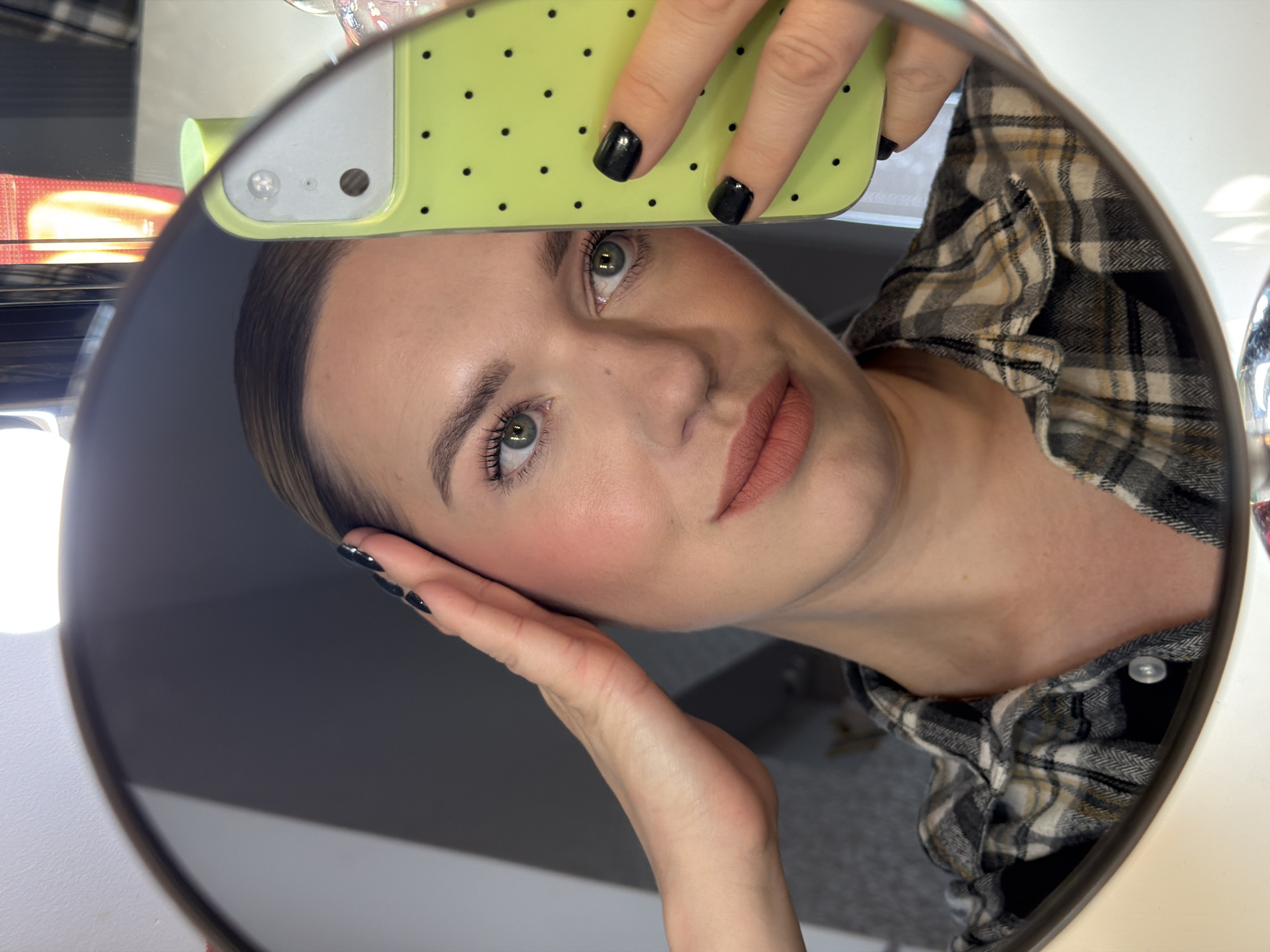 A woman is reflected in a round mirror. She is holding a green smartphone with black dots on its case. Her fingernails are painted black. She is wearing a plaid shirt and has makeup on, including blush and lipstick. The location is likely not in Dreiliņi, Latvia, as the image does not contain any discernible location-specific details.