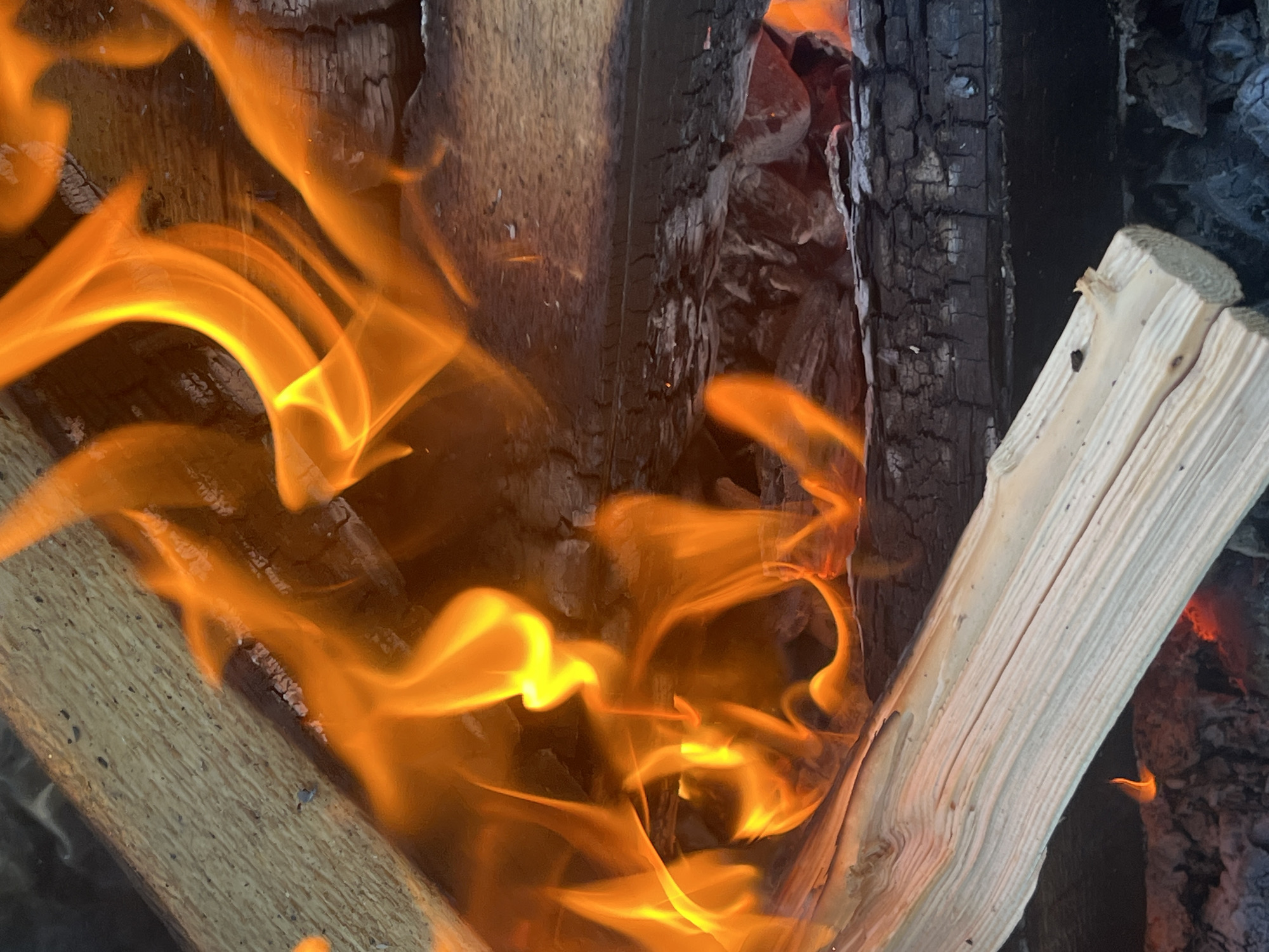 The image is a close-up, high-angle shot of a campfire with brightly burning orange flames engulfing logs. The logs are stacked closely together, with some appearing charred and dark, while others are lighter in color and show distinct wood grain. The fire casts a warm glow on the surrounding wood and embers. There are no people visible in the image. The setting appears to be outdoors, with no identifiable landmarks to pinpoint a specific location in Il'ino, Russia. The focus is entirely on the fire and the burning wood.