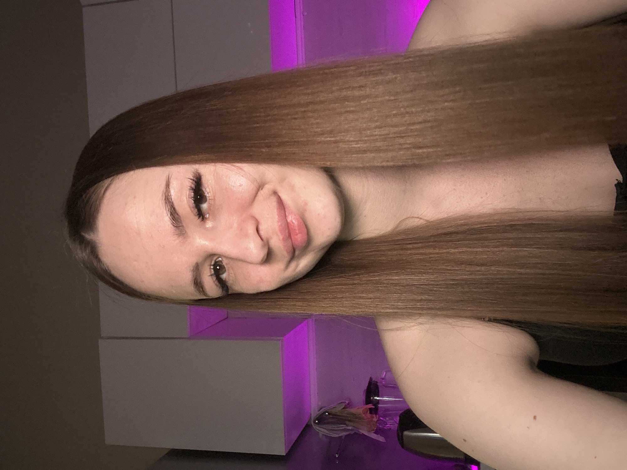 A woman with long, straight brown hair is positioned with her head slightly tilted towards the viewer. The background features a white shelf with a purple light emanating from behind it. Part of her shoulder is visible in the lower right corner. The location is Unknown, Unknown.