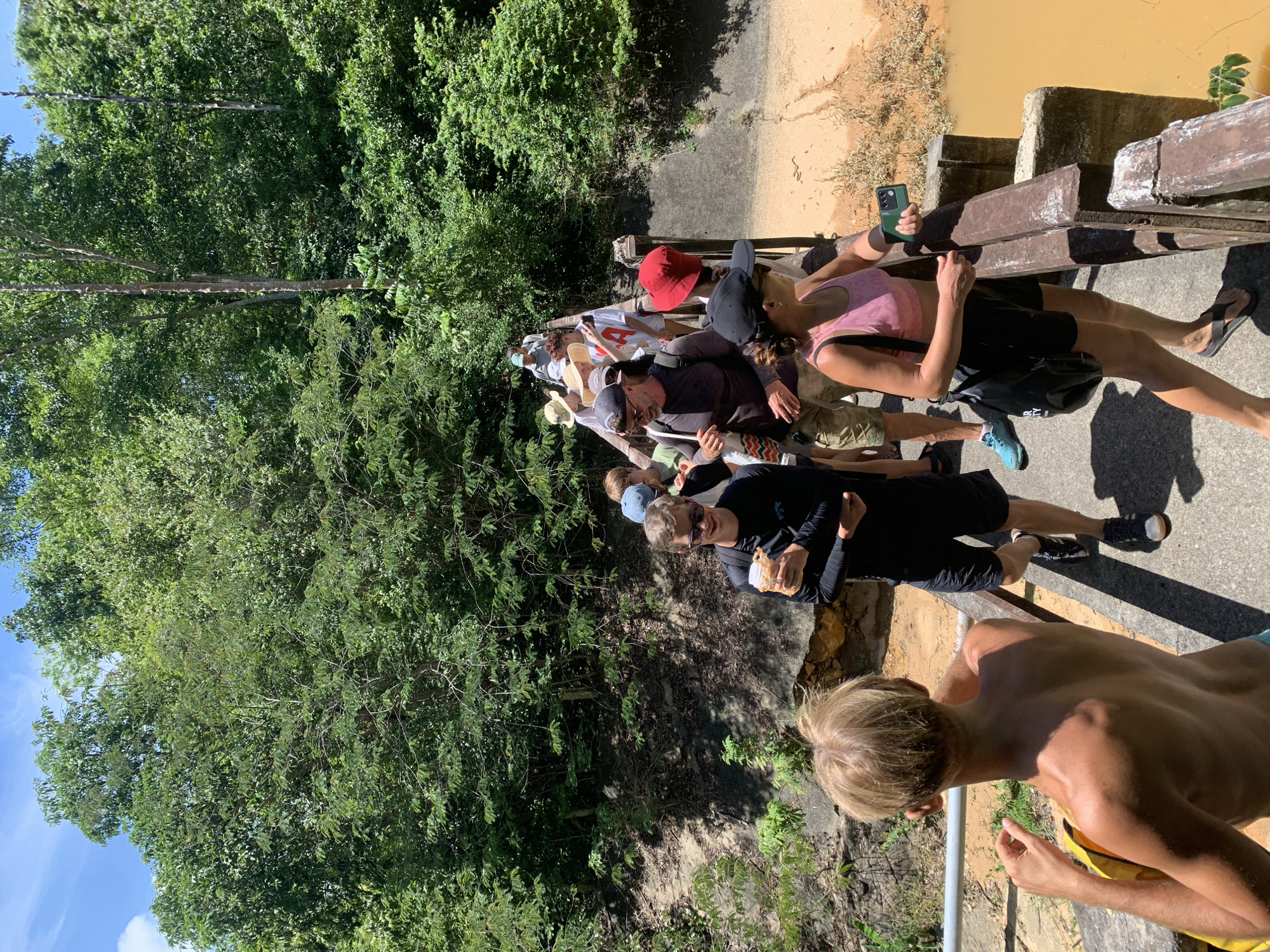 The image shows a group of tourists walking on a wooden bridge over a muddy, reddish-brown body of water, likely a river or canal, surrounded by lush green vegetation and trees. The sky is blue with some white clouds. The tourists are dressed in casual, warm-weather clothing, including shorts, t-shirts, and hats. Some are carrying bags or holding drinks. The bridge appears to be part of a nature trail or park. One tourist in the foreground on the right is holding a green smartphone. The overall scene suggests an outdoor excursion in a tropical environment. Without additional context, the specific location in Ko Samui District, Thailand cannot be definitively determined from the image alone.
