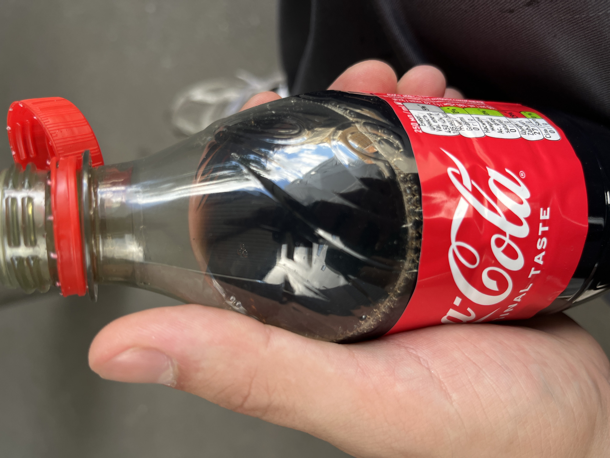 A hand holds a plastic bottle of Coca-Cola. The bottle is partially filled with dark liquid and has a red cap. The label is predominantly red and white, with the Coca-Cola logo and nutritional information visible. The bottle is held at an angle, with reflections visible on its surface. The background is blurry and appears to be outdoors, with a dark, textured surface and hints of natural light. The image was taken in Kyiv, Ukraine.