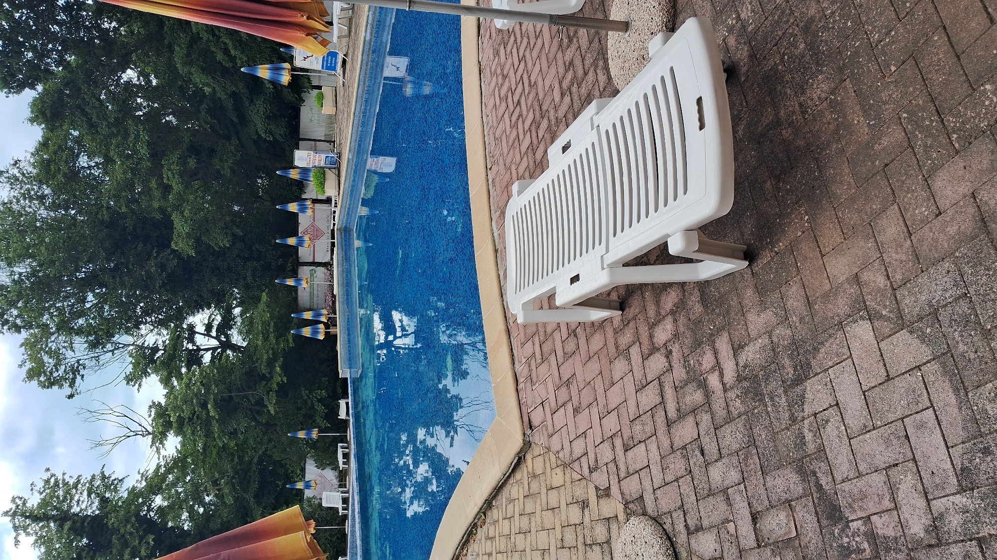 The image depicts an outdoor swimming pool area, likely located in Varna, Bulgaria, as indicated by external information. The pool is filled with blue water and is surrounded by a beige tiled border. The pool deck is paved with reddish-brown bricks arranged in a herringbone pattern. A white plastic lounge chair is positioned prominently in the foreground on the brick paving, angled towards the right of the frame. Several beach umbrellas with orange, yellow, and blue stripes are visible along the perimeter of the pool, some open and others closed. In the background, a dense line of green trees forms a natural backdrop. Behind the pool, a low wall is visible with various signs and posters, suggesting a commercial or recreational establishment. There are no people visible in the image. The sky is overcast with a few white clouds. The overall impression is that of a quiet, perhaps slightly weathered, outdoor pool setting.