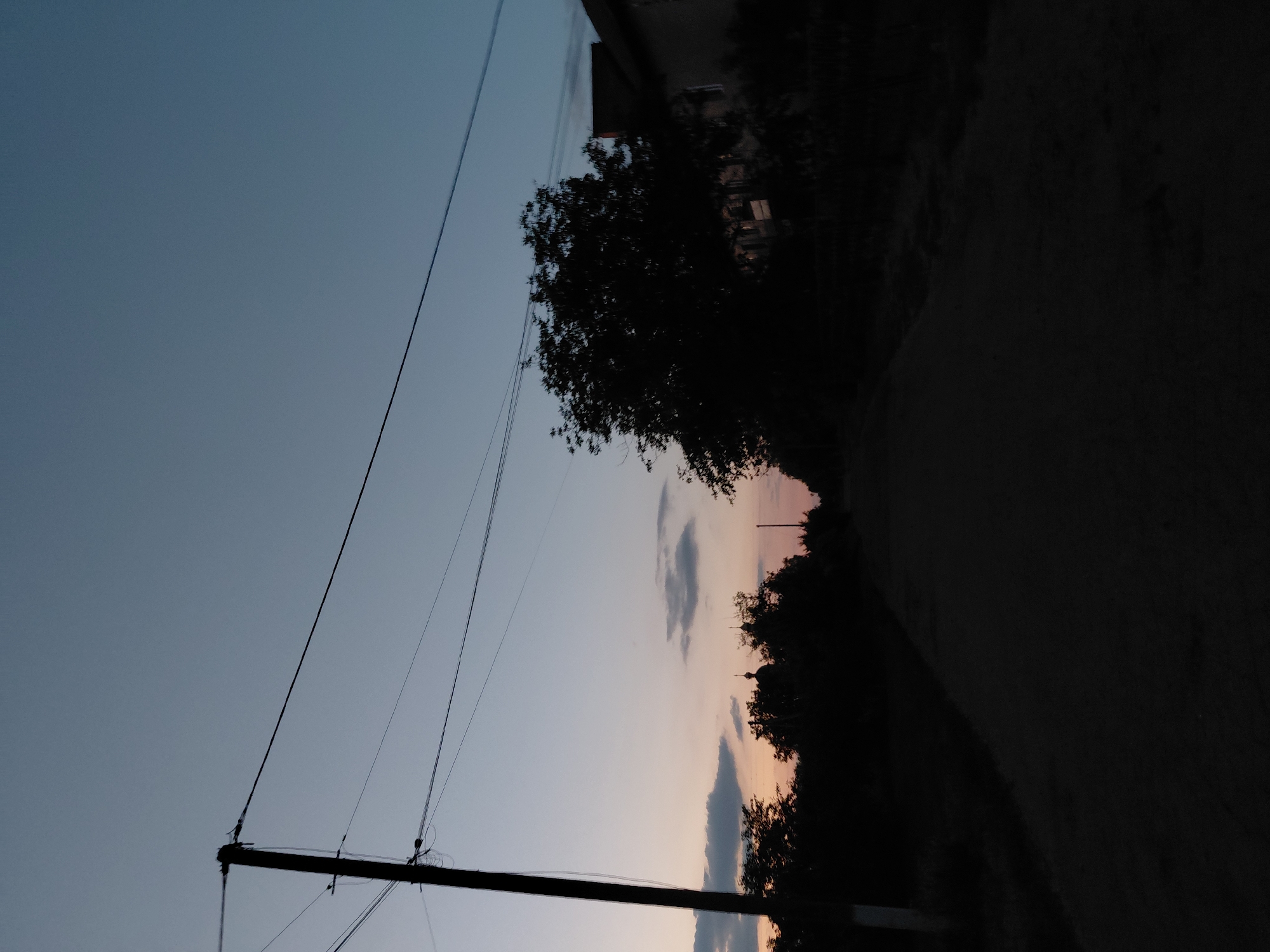 The image shows a twilight scene on a dirt road in Ustyvytsia, Ukraine. A wooden utility pole with electrical wires stands on the left side of the road. The sky is a gradient of pale blue and pink, with scattered clouds. Silhouetted against the sky are trees, a building on the right, and a church dome in the distance. The road itself is dark and empty, with no people or vehicles visible. The overall impression is one of quiet and dusk in a rural setting.