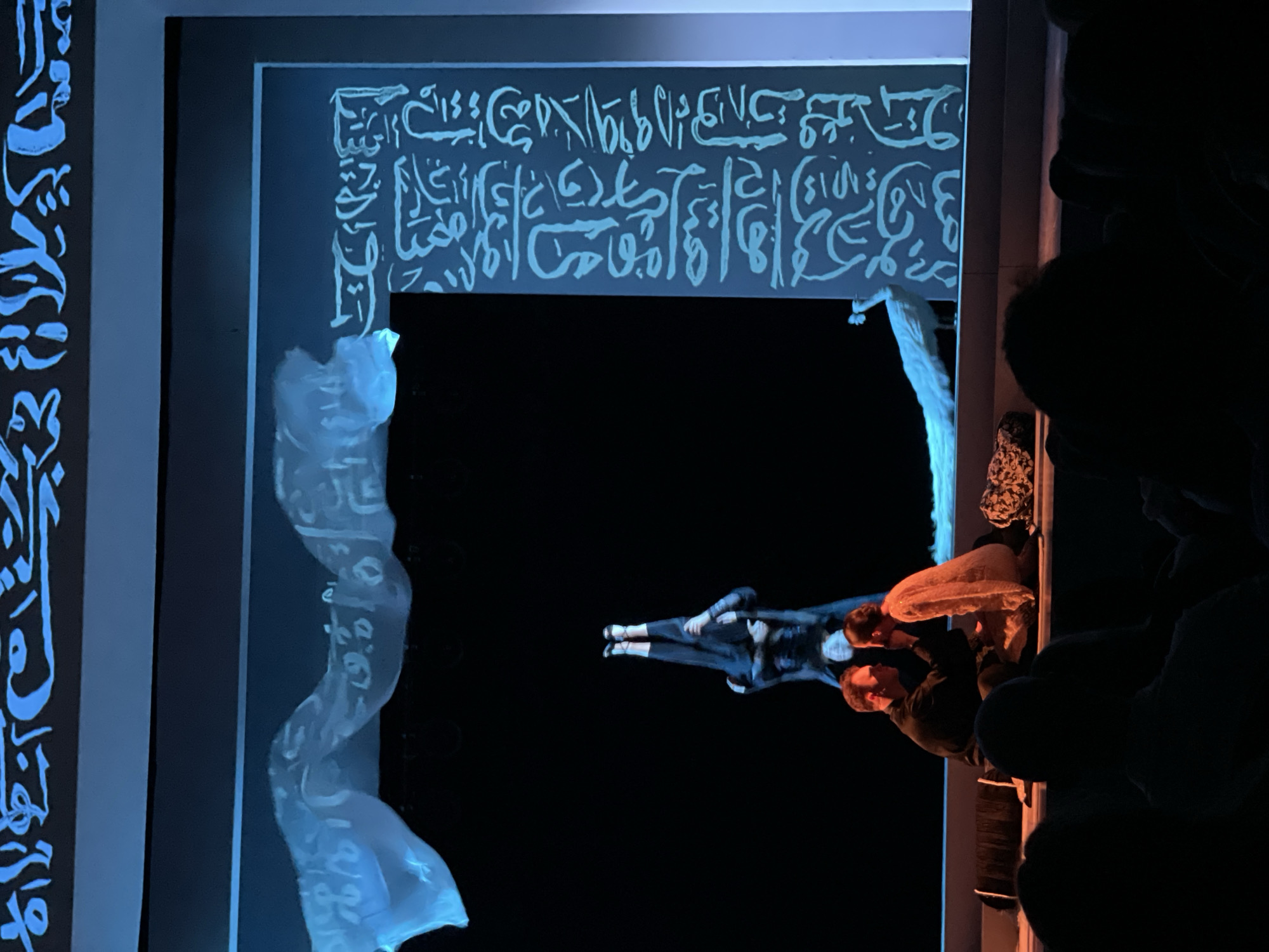 The image captures a performance within a theater setting, possibly in Krasnoyarsk, Russia. Two performers, a male and a female, are centrally positioned against a black backdrop, suggesting an absence of traditional scenery. The male is dressed in black attire, while the female appears to be wearing a light-colored garment. Above them is a projection of white Arabic script on a dark blue rectangular canvas, which appears to be affixed to the wall. To the left, a sheer white fabric with projected Arabic script drapes vertically, resembling a curtain or theatrical prop. The scene is lit with blue and white lighting, which accentuates the projection and the performers. Audience members are visible in the darkness on the right, suggesting the performance is actively being viewed.