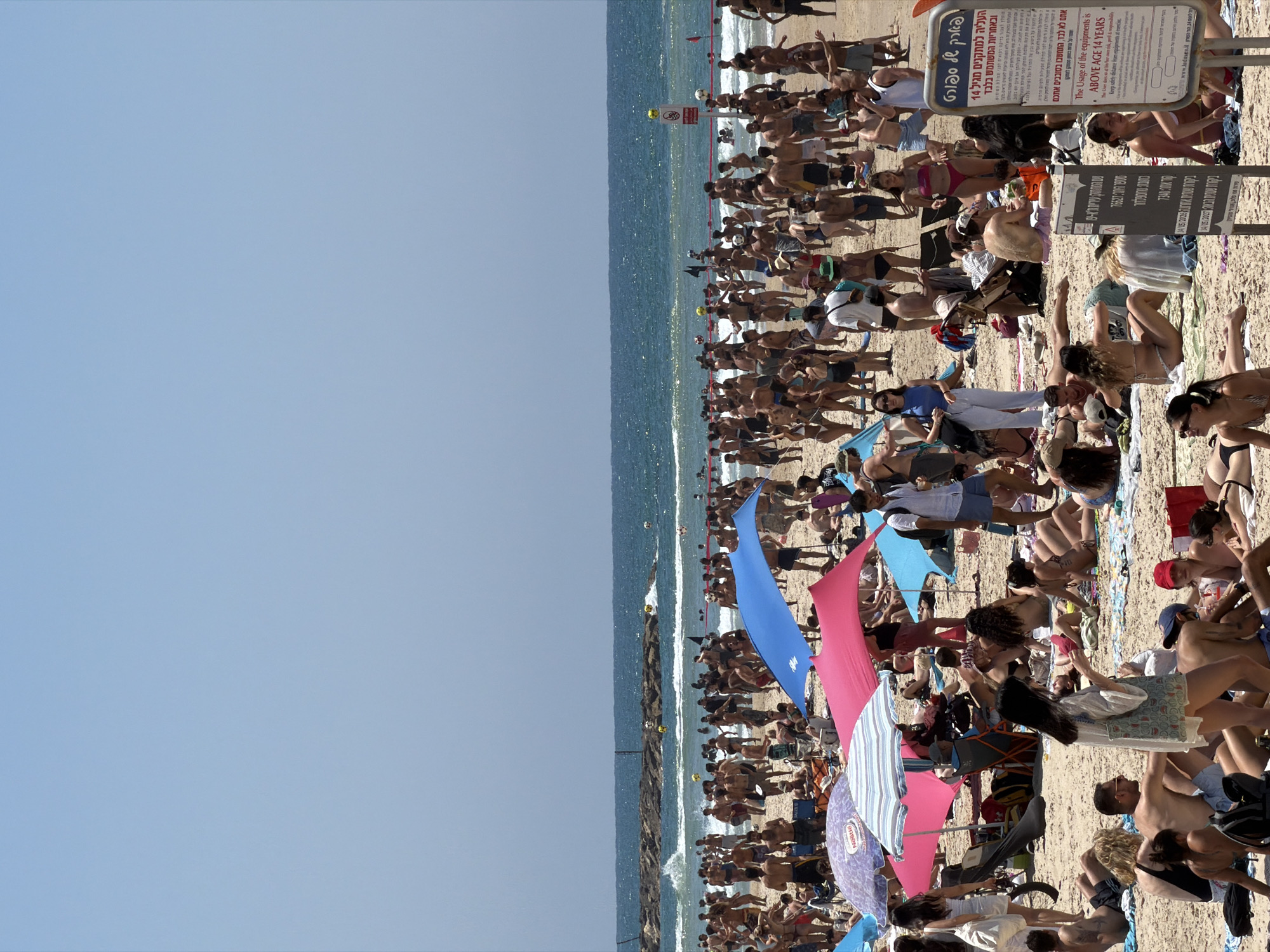 A beach scene in Tel Aviv, Israel, shows a crowded beach with numerous people sunbathing, swimming, and relaxing. The sky is clear and blue. The ocean is visible in the background. There are signs posted with safety information and regulations. Umbrellas provide shade for beachgoers.