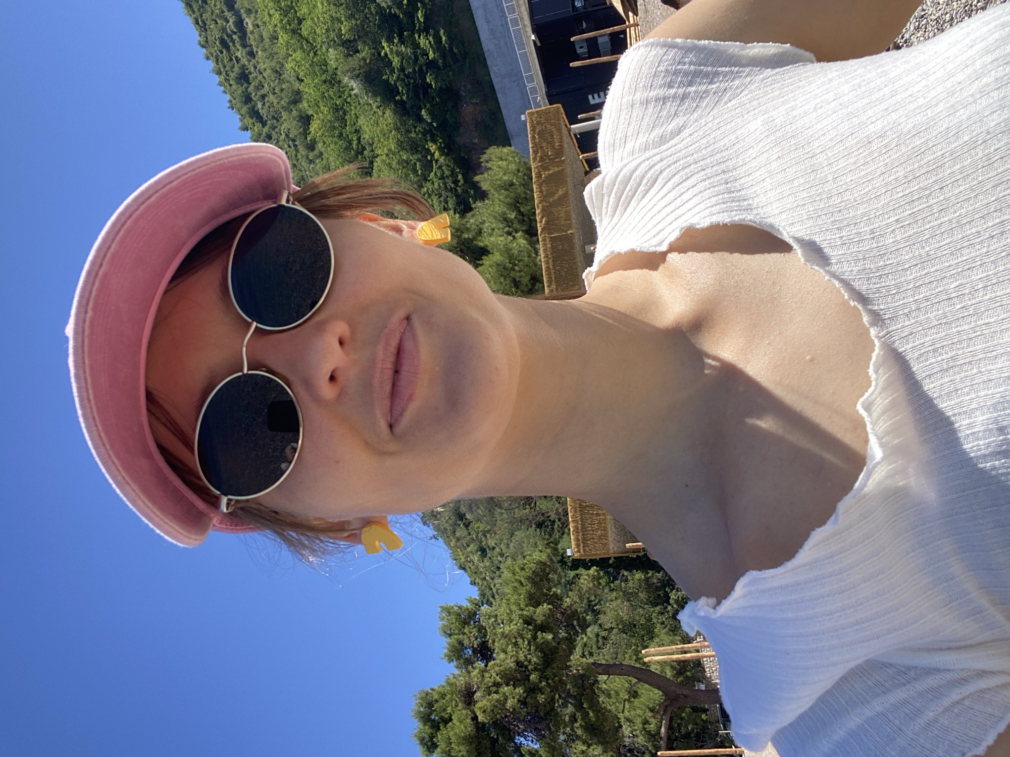 A selfie of a woman wearing a pink hat and black sunglasses. She is wearing a white ribbed top. In the background, there is a tree and a bright blue sky. Based on the vegetation and architecture, the image likely was taken in the city of Budva, Montenegro.
