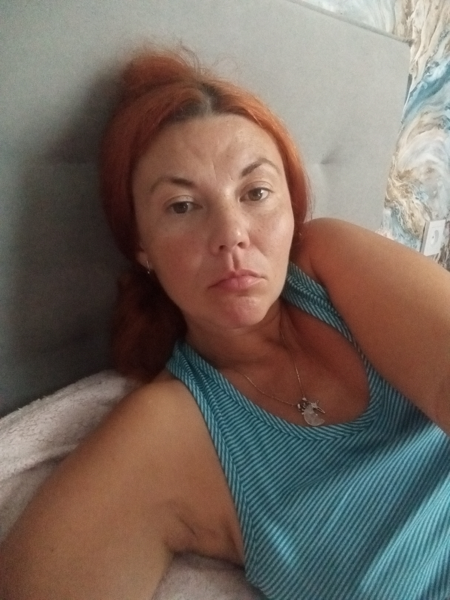 A medium shot of a woman lying on her side, looking at the camera. She has red hair pulled back and is wearing a blue and white striped tank top. She has a necklace with a pendant. The background appears to be a wall with abstract blue and white wallpaper and a gray headboard or wall. The lighting is soft and appears to be natural, coming from the side.