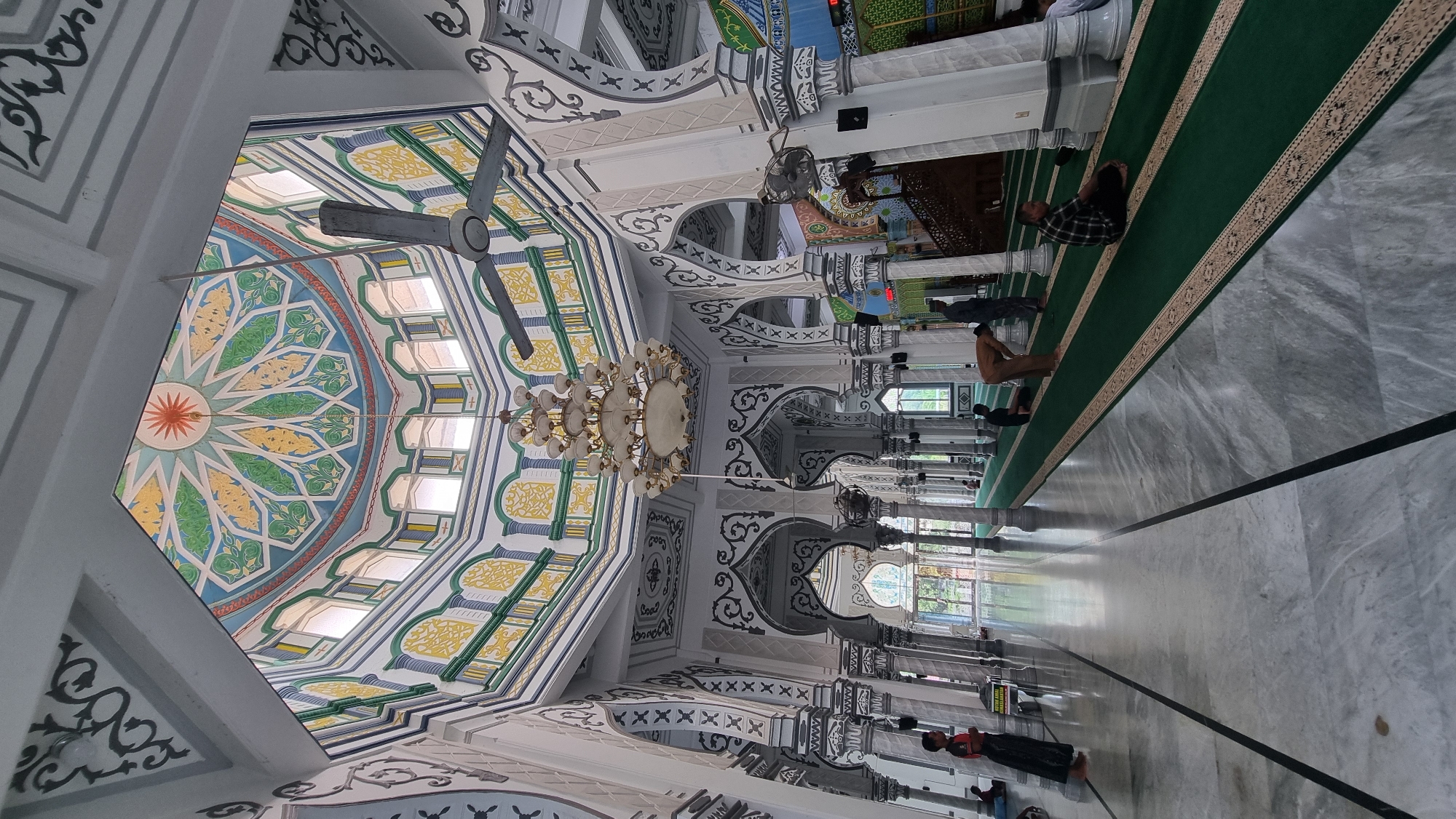 The image shows the interior of a mosque, likely the Masjid Al-Munawarah in Keudekruenggeukueh, Indonesia, as indicated by its distinctive architecture. The central focus is the high, ornate dome ceiling, decorated with a geometric pattern in blue, green, and yellow, with a sun-like motif at its apex. A large, multi-tiered chandelier hangs from the center of the dome.
The space features tall, fluted columns with intricate grey and white decorative elements supporting arches. A ceiling fan is visible near the dome. The floor is polished marble with dark grey lines, and green prayer mats with a decorative border are laid out in rows.
Several individuals are present in the mosque. In the foreground, on the left, a young person stands near a pillar, wearing a dark shirt and a long skirt or sarong. Further into the mosque, on the green mats, several men are engaged in prayer or seated. One man in a light brown garment is in a bowing position, while others are in a kneeling or seated posture. Another man in a plaid shirt is seated facing to the right. The overall environment suggests a serene and spiritual atmosphere during prayer time. The architecture appears well-maintained, with clean lines and vibrant decorative details contributing to the aesthetic appeal of the prayer hall. Natural light filters in from windows along the upper sections of the walls.